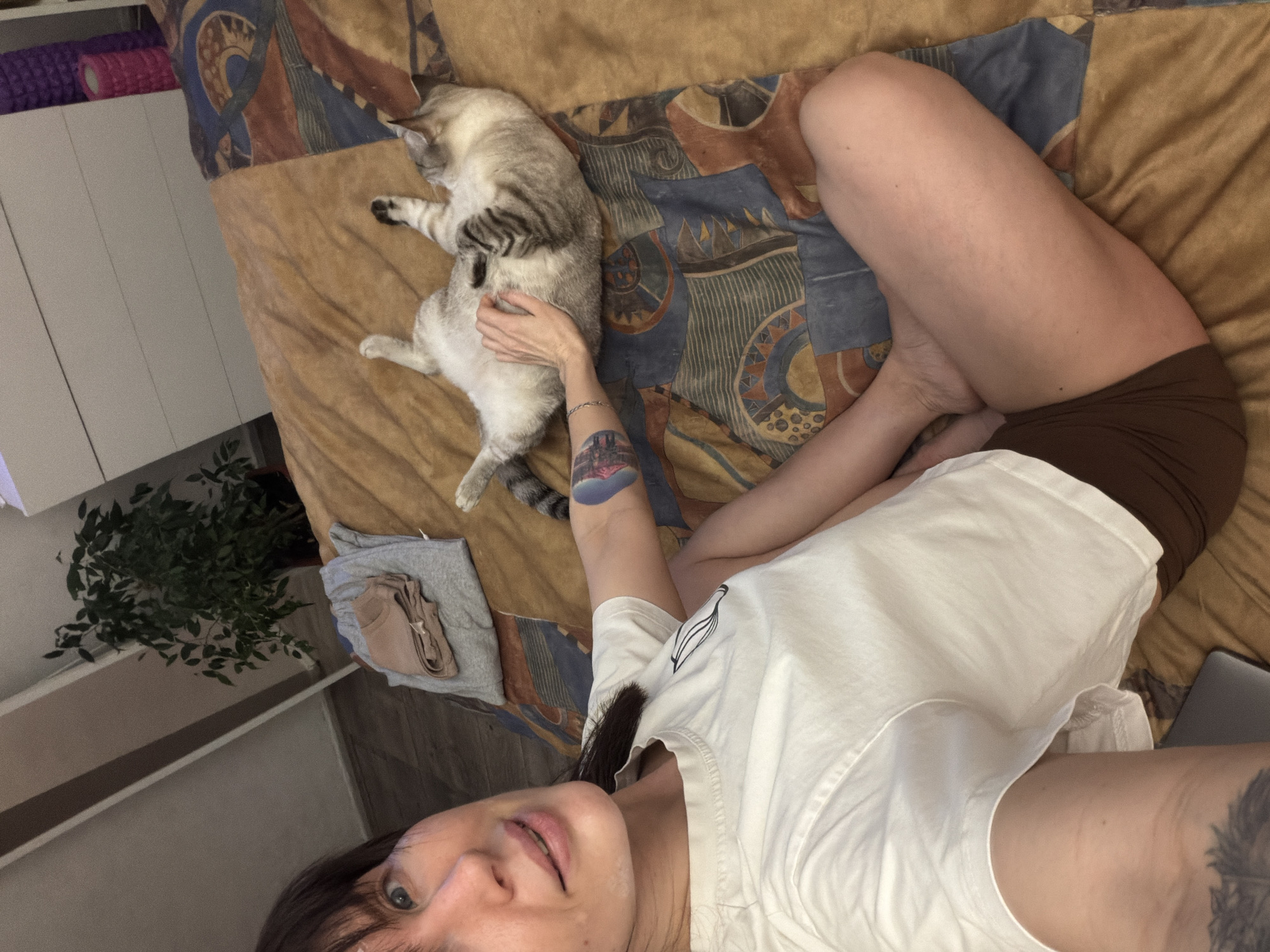 The image, captured from a low-angle selfie perspective, depicts a person relaxing on a bed with a cat. The female subject, with dark hair and bangs, looks towards the camera with a neutral to slightly smiling expression. She is dressed in a white t-shirt and brown shorts. A colorful tattoo, possibly a landscape scene, is visible on her left forearm, along with a silver bracelet. A darker, intricate tattoo is partially visible on her right upper arm.
Beside her, a light-colored, striped tabby cat lies on its back or side, appearing very relaxed with its belly exposed. The person's hand is gently placed on the cat's stomach, indicating she is petting it.
They are positioned on a bed covered with a quilted blanket featuring an abstract, geometric pattern in hues of brown, blue, and orange. A closed gray laptop is visible on the blanket near the person's legs. A small stack of folded clothes, including gray and beige items, rests on the bed.
In the background, a white cabinet or shelf unit is visible, topped with purple and pink foam rollers. A lush green houseplant is situated on the floor next to the bed. The scene conveys a calm, domestic atmosphere, likely indoors in a bedroom during daytime. No discernible text is present in the image.
