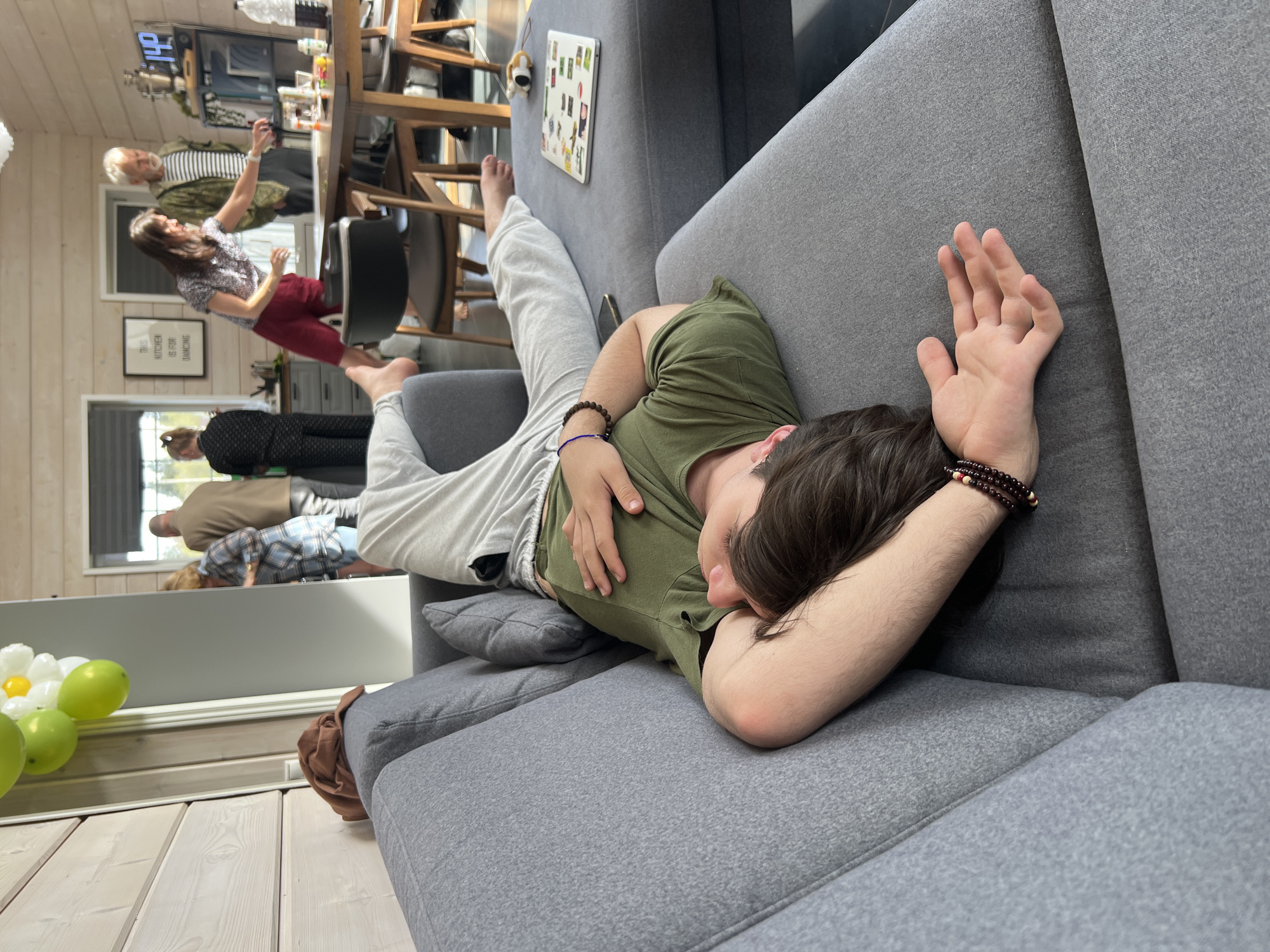 A young male is asleep on a grey couch with his head resting on his arm. He is wearing a green t-shirt and light grey sweatpants. He has brown hair and is wearing bracelets on both wrists. In the background, reflected in a mirror, are several adults standing in a kitchen and dining area. There is a table with chairs, and a woman in a floral shirt and red pants appears to be dancing. The location is Svetloe, Russia.