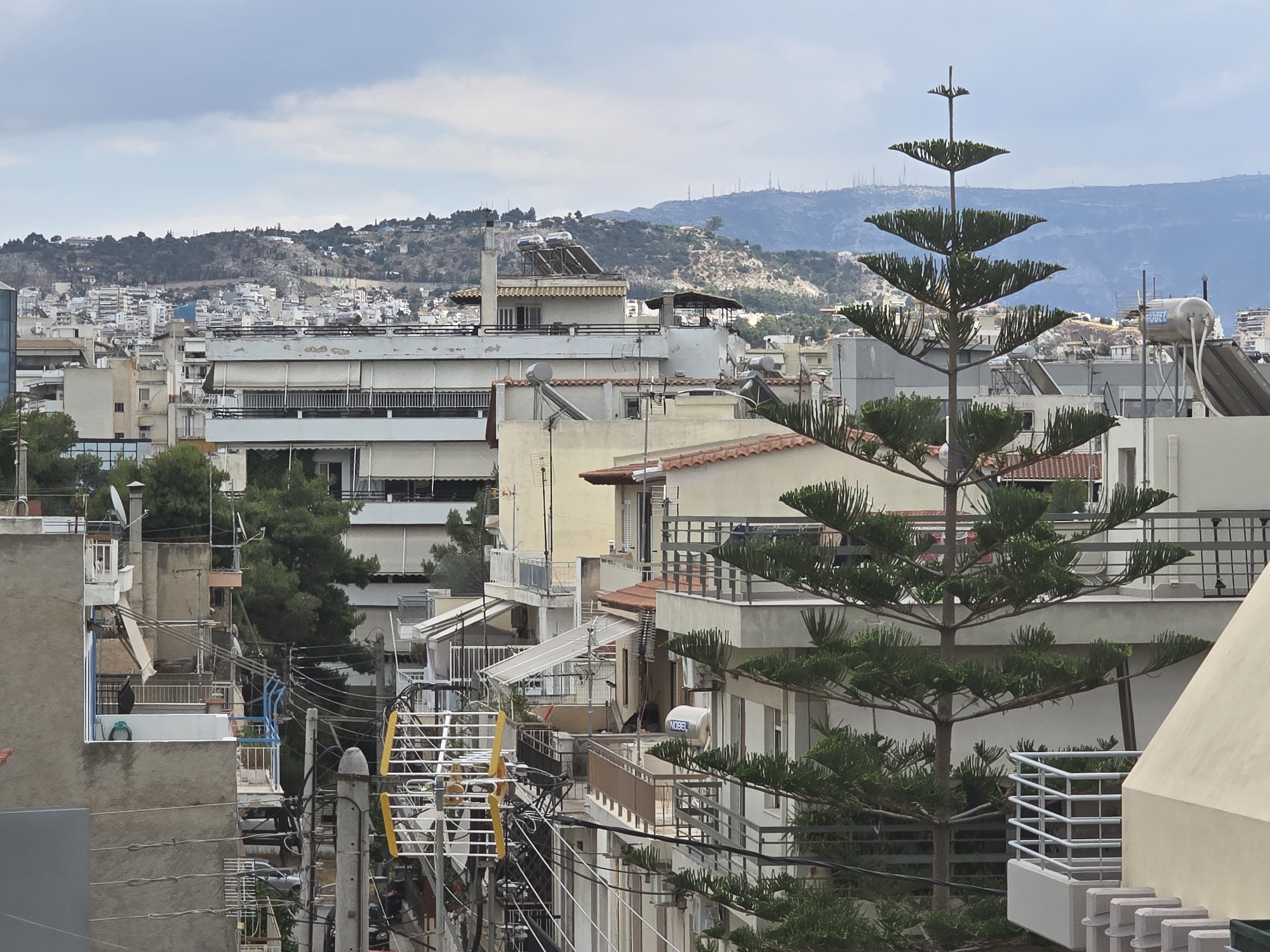 This media file captures an elevated view of a dense urban and suburban landscape in Agii Anargiri, Greece, on an overcast day. The scene is dominated by numerous multi-story residential buildings, primarily light-colored (white, beige, and light grey), featuring multiple balconies with metal or concrete railings. Many rooftops are flat and equipped with common Greek amenities such as solar water heaters – one white cylindrical tank prominently displays the brand "NOBEL" – and various antennas. Some buildings also feature traditional terracotta-tiled roofs.
A tall, distinctive coniferous tree with tiered branches stands prominently in the mid-ground on the right side, adding a natural element to the built environment. Throughout the mid-ground and left, a complex web of overhead power lines and utility poles traverses the scene, illustrating typical urban infrastructure.
In the background, a range of hills and low mountains stretches across the horizon, appearing rugged and partially covered with sparse vegetation. These hills are also dotted with smaller structures and what seem to be communication towers or antennas on their peaks. The sky is uniformly grey and cloudy, indicating an overcast day without direct sunlight. No people are visible, and there is no discernible activity or event taking place, presenting a static snapshot of an everyday cityscape.