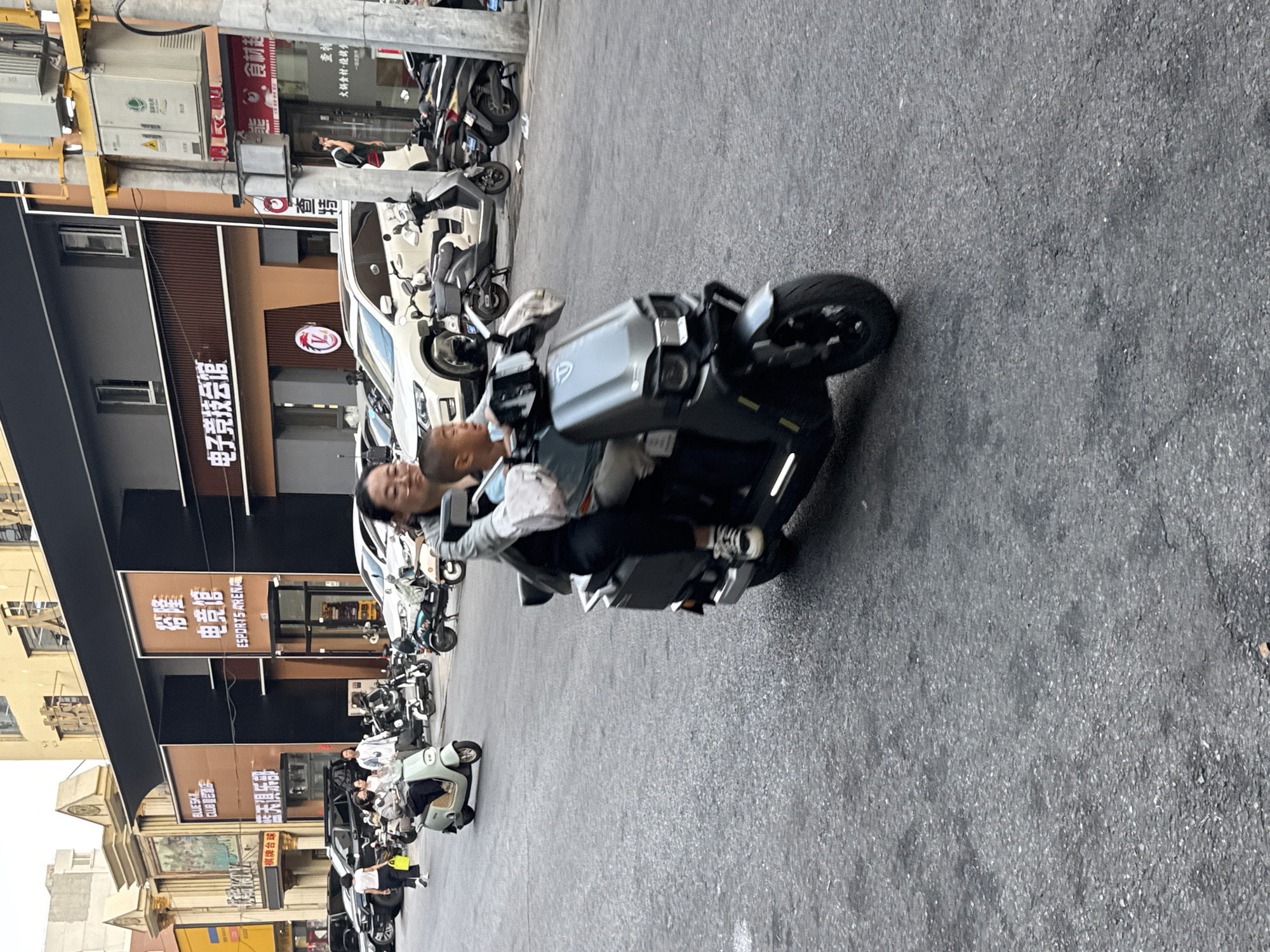 In Guta, China, a woman and a young boy are riding a grey electric scooter on a paved street. The woman is driving, and the boy is seated behind her. They are both wearing casual clothing. The scooter is moving forward, and the background shows a street scene with buildings, other scooters, and parked cars. One of the buildings has signage in Chinese characters. The overall impression is a typical urban street scene.