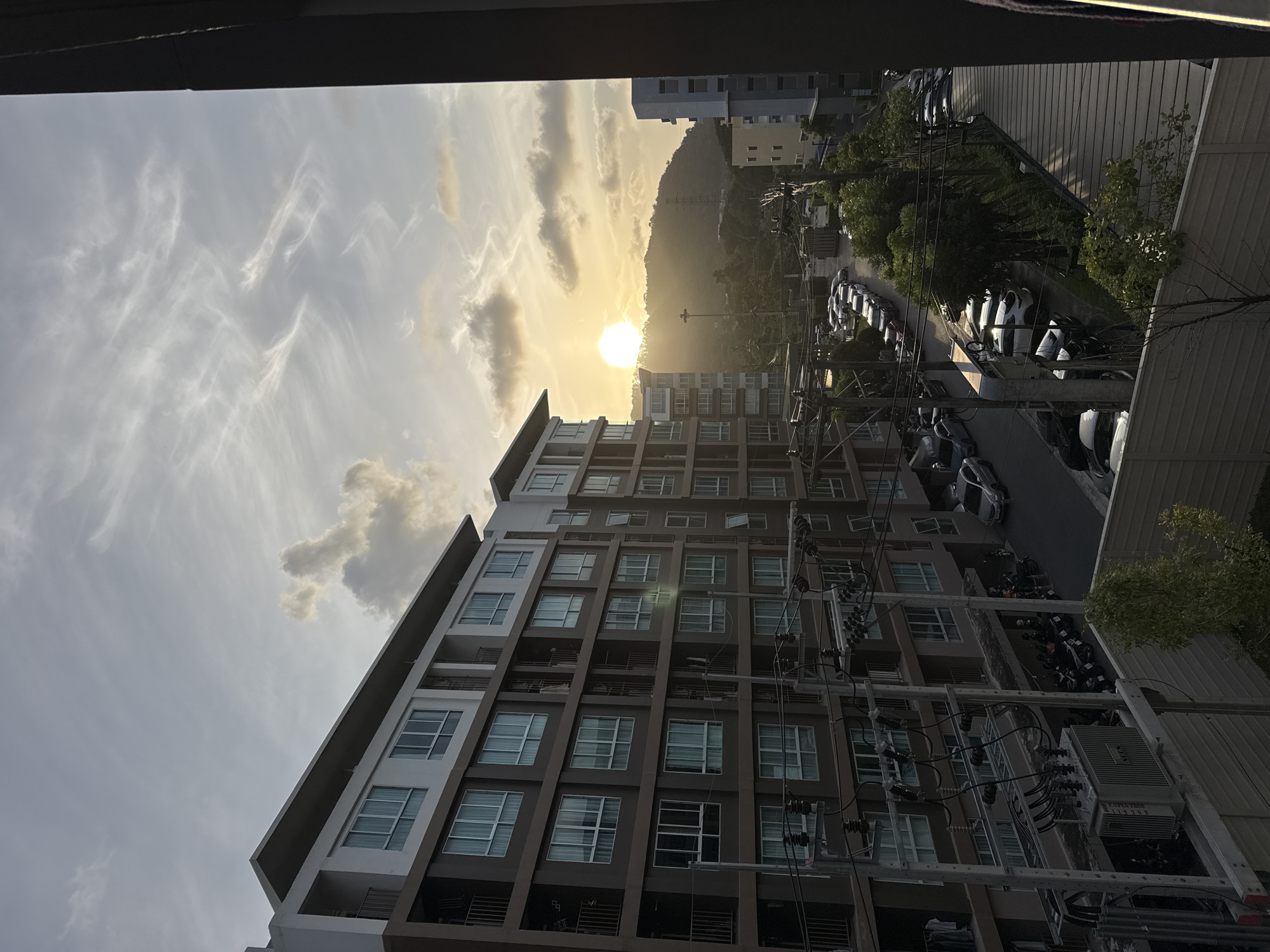 A dusk view from an elevated perspective in Kathu, Thailand, captures a residential complex and its surroundings. The foreground features an electrical transformer and power lines connected to a multi-story building. To the left, a building with a brown facade and numerous windows stretches across the frame, with balconies visible. Beyond this, a street is lined with parked cars and motorcycles. In the background, another building, lighter in color, is partially visible, and further beyond that, a hill is silhouetted against a sunset sky. The sun, a bright orb, is low on the horizon, casting a warm glow and creating a hazy atmosphere. Wispy clouds stretch across the sky. The scene appears to be in a densely populated urban or suburban area.