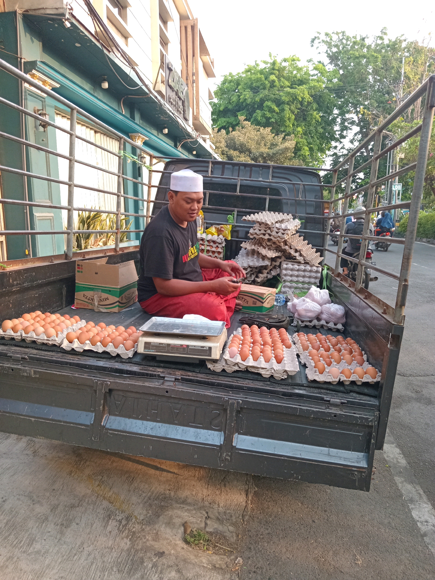 In Randuagung, Indonesia, a man wearing a white peci cap and a black t-shirt sits on the bed of a black pickup truck, which is loaded with numerous trays of brown eggs. He is wearing a red sarong. In front of him, on the truck bed, is a digital scale and several packs of eggs. Behind him, more egg cartons are stacked high, along with several plastic bags and a cardboard box. The truck is parked on a concrete surface next to a street where a few motorcycles are visible in the background. The scene appears to be set up for selling eggs, possibly at a market or roadside stall. The building in the background is light-colored with teal trim.