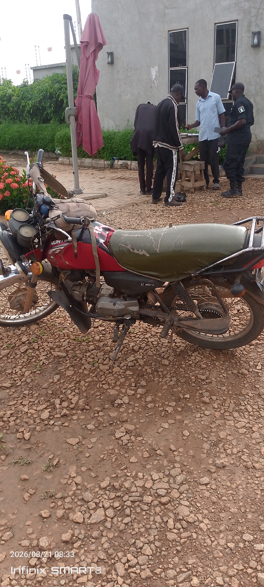 In Kakau, Nigeria, a red and black motorcycle with a damaged olive green seat sits on a gravel surface. Behind the motorcycle, a pink umbrella is tied to a pole. Further in the background, four men are gathered near a building with a concrete facade and large windows. One man, wearing a light blue shirt and dark pants, is speaking to another man in a black and white tracksuit. Two other men are dressed in black uniforms with "NIGERIA POLICE" visible on one's arm. The ground is uneven and covered with loose stones.