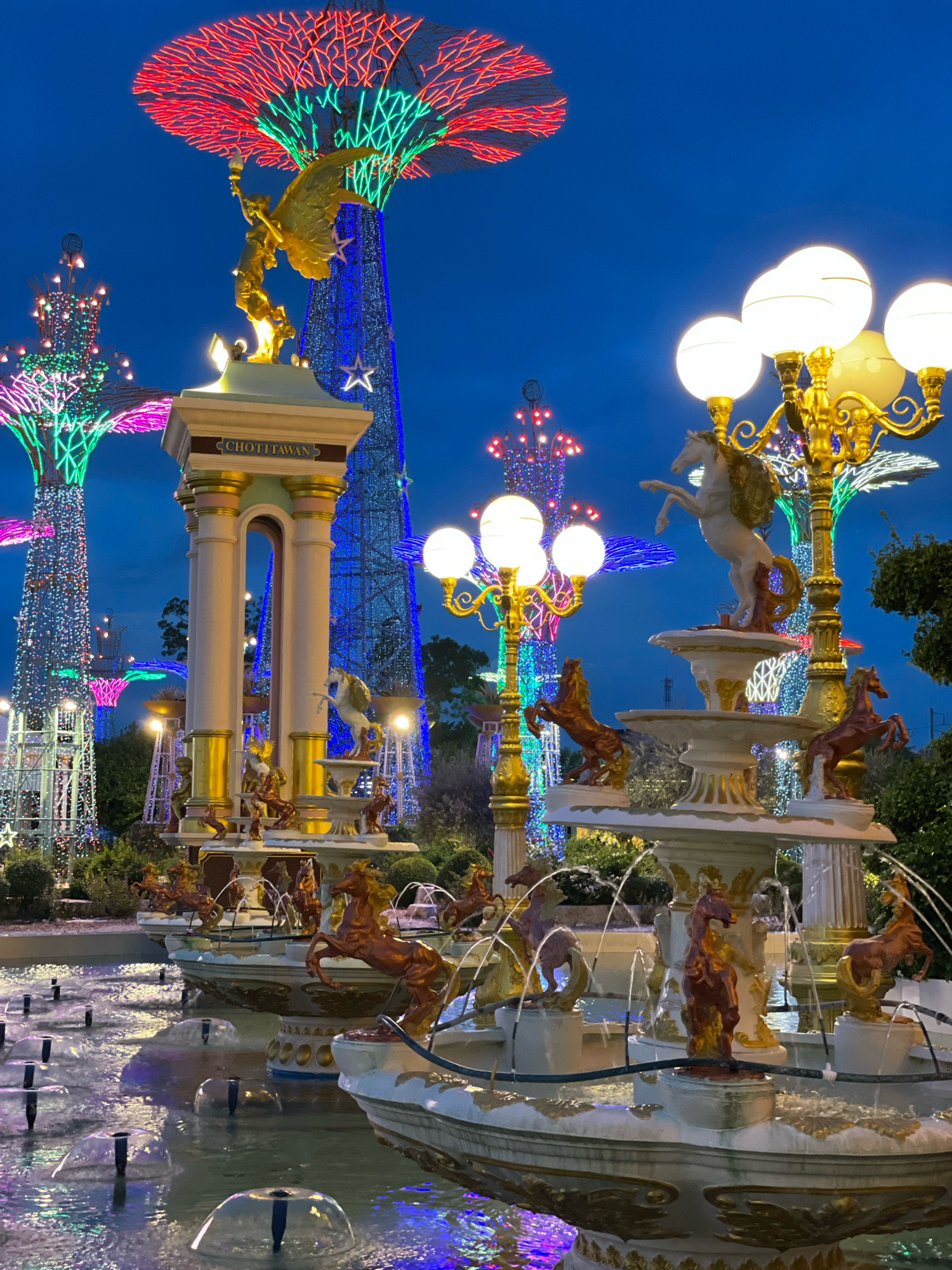 The image depicts a nighttime scene at the Pattaya City Light Festival in Pattaya, Thailand. The foreground features a multi-tiered fountain with ornate sculptures of horses and human figures, with water jets creating a shimmering effect. Behind the fountain, a classical-style archway with gilded accents stands prominently, bearing the inscription "CHOTITAWAN." Further in the background, elaborately decorated "trees of light" with colorful LED lights in red, green, blue, and purple illuminate the dark sky, creating a festive atmosphere. Ornate lampposts with multiple globe-shaped lights and gilded detailing are also visible. The overall scene is characterized by vibrant artificial lighting and elaborate decorative structures, indicative of a special event or attraction.
