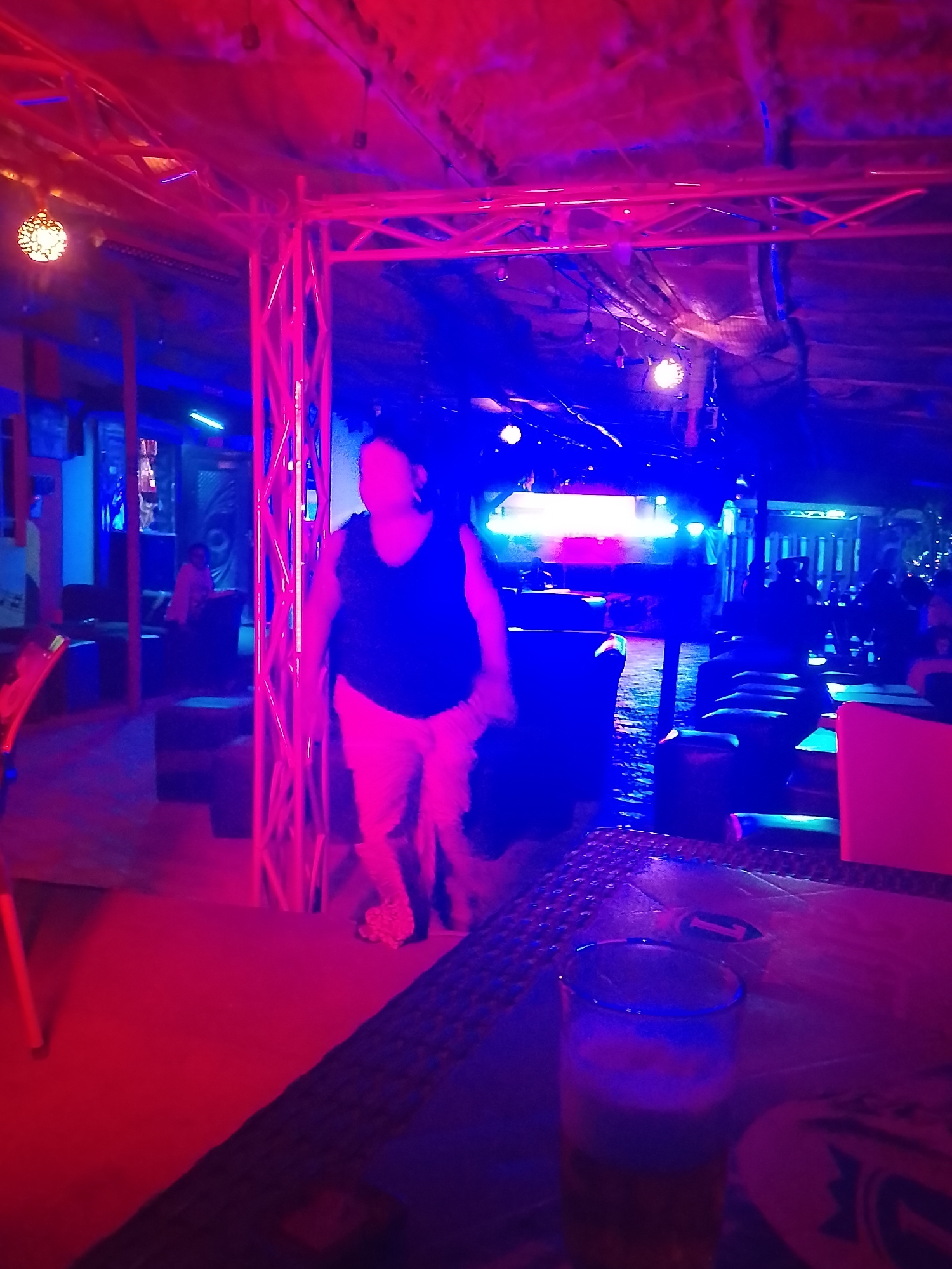 This image captures an indoor nightlife scene, likely a bar or club, enveloped in vibrant artificial lighting. The dominant colors are intense red and blue, which cast dramatic hues across the entire environment, making specific details and true colors difficult to discern.
In the mid-ground, slightly to the left, a person is depicted in motion, walking towards the right. They are wearing a dark sleeveless top and light-colored pants. Their figure is heavily illuminated by the colored lights, rendering their facial features indistinct and giving them a somewhat blurred appearance. Several other patrons are visible in the background, seated or standing, also bathed in the club's lighting, indicating a social gathering.
The foreground on the right shows a table with a dark, patterned surface. On it, a tall glass containing liquid reflects the blue and red light. A small white object, possibly a coaster, is also on the table. To the left, a portion of a red support structure or furniture leg is visible, alongside dark, cushioned seating areas.
Above, exposed metal truss structures, likely for lighting and sound equipment, span the ceiling. The ceiling itself appears to be covered with a dark, textured, or possibly thatched material. A decorative hanging lamp with a patterned globe emits a warmer light on the upper left side. In the deeper background, a bright, rectangular light source stands out, further illuminating the space.
The scene depicts an active, social environment at night. No discernible text is visible within the image. The setting suggests a lively entertainment venue, consistent with nightlife in a location such as Yelwa, Nigeria.
