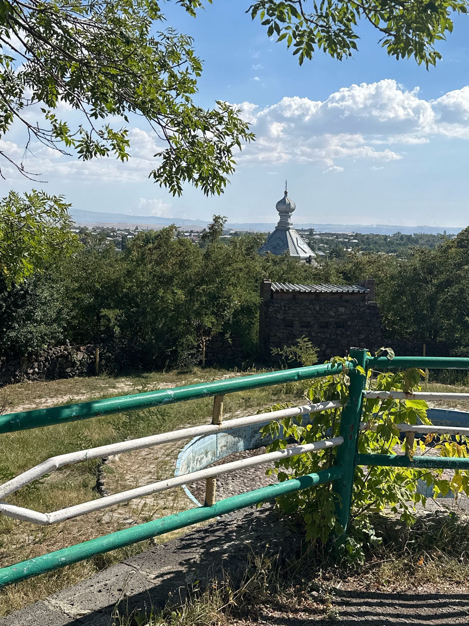 A view of the city of Gyumri, Armenia, shows a landscape dotted with buildings and lush greenery under a bright blue sky with fluffy white clouds. In the foreground, a section of a green and white railing partially obscures a paved area and a small stone structure with a corrugated metal roof. Beyond this, trees and foliage create a dense canopy, with a distinctive domed building, possibly a church, visible in the mid-ground. The distant horizon reveals a sprawling urban area and rolling hills. The image captures a sunny day, with shadows cast on the ground in the foreground, suggesting the sun is high in the sky.