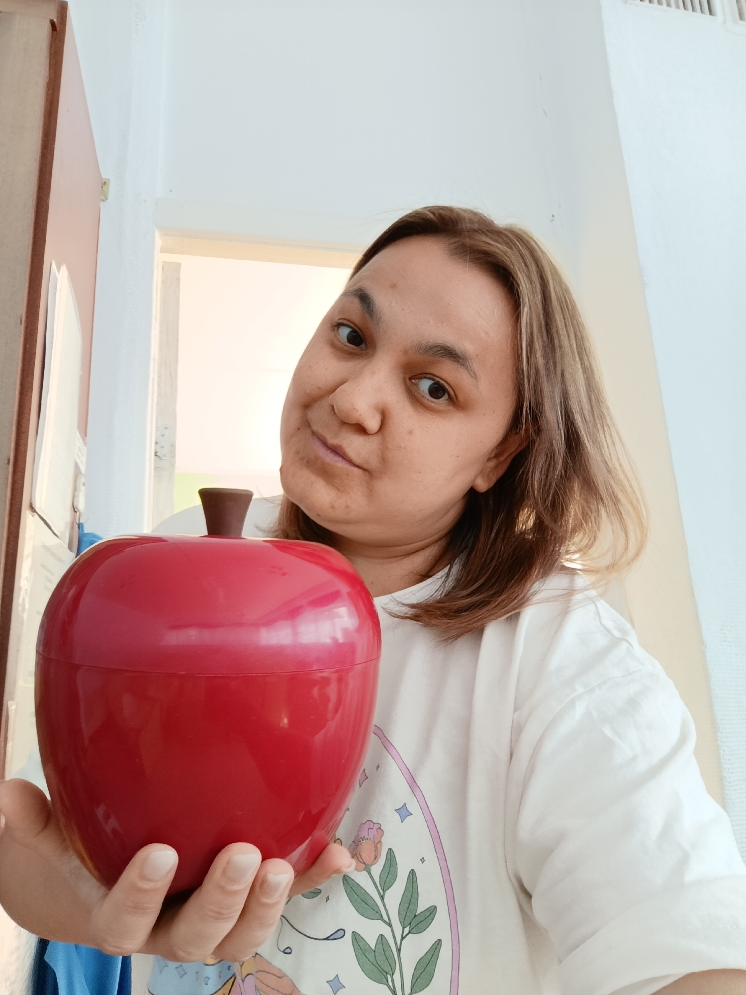A woman holds a large, shiny red apple-shaped container. The woman has medium-length brown hair and is wearing a white t-shirt with a floral design. The background shows a doorway and white walls. The location is likely an interior space, possibly a home. There is no information confirming location within the city of Chaikovsky, Russia.