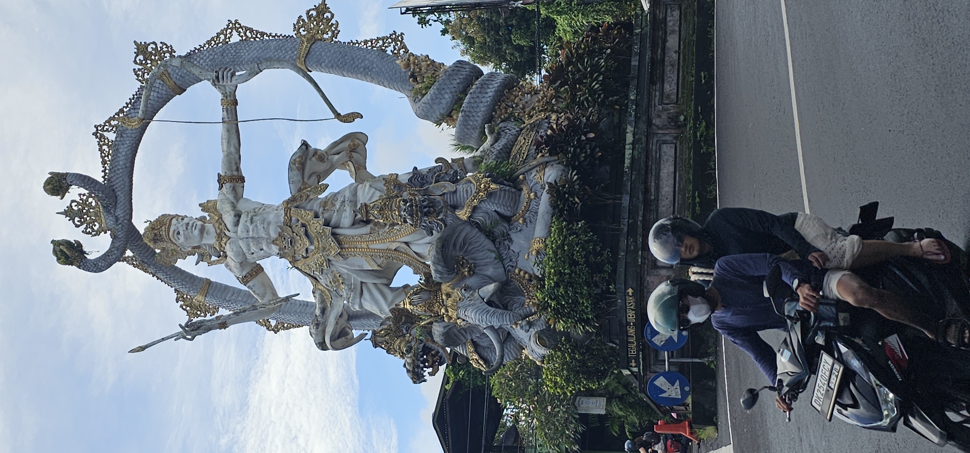 The image captures a scene in Ubud District, Indonesia, featuring the Arjuna Wijaya statue at a roadside. The statue, predominantly white with gold embellishments, depicts a figure in a heroic pose, holding a bow and arrow. A multi-headed serpent-like creature is entwined around the statue. Two individuals on a parked motorcycle are visible on the right side of the frame, next to the street. One is sitting on the motorbike wearing a helmet and face mask, and the other laying behind, also wearing a helmet. A traffic sign displaying directions toward "TEGALLALANG - DENPASAR" is placed beside the road. The environment includes tropical foliage and a partially visible structure. The sky is partly cloudy.
