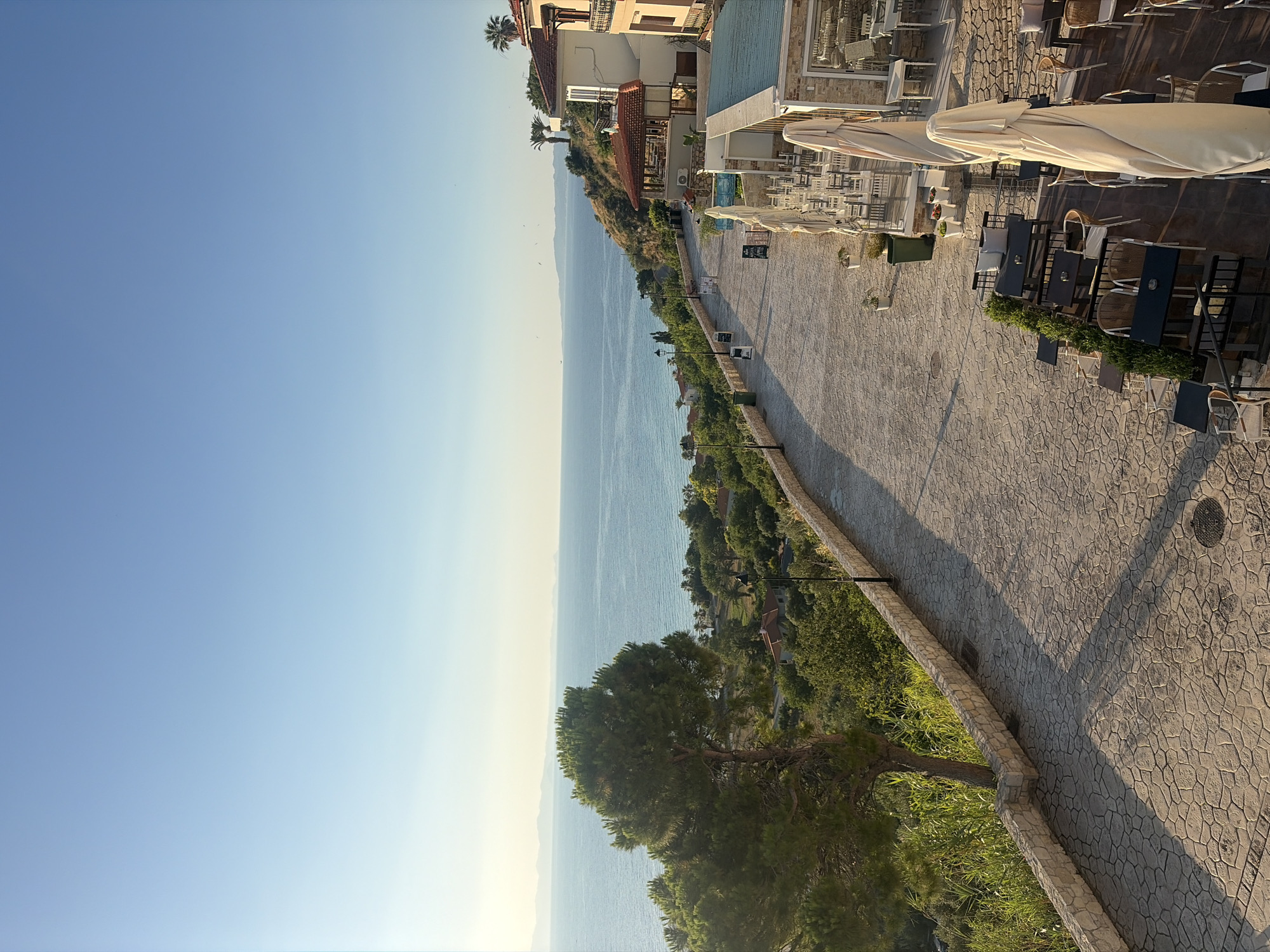 The image depicts a scene in Kassandra Municipal Unit, Greece, looking out over the Aegean Sea. A stone-paved walkway lines the edge of a cliff, with lush green foliage and trees descending towards the sea. The walkway is bordered by a low stone wall on one side.
Along the right side of the walkway, several outdoor seating areas for cafes or restaurants are visible. These areas feature tables and chairs, some covered by large, light-colored umbrellas. Buildings, typical of a Mediterranean coastal town, are situated behind these seating areas, characterized by terracotta roofs and light-colored facades.
The sea is a prominent feature, stretching to the horizon under a clear, pale blue sky. Distant landmasses are faintly visible on the horizon. The sun appears to be low in the sky, casting long shadows across the walkway and creating a warm, golden light.
No people are clearly visible in the immediate foreground or midground of the image. The overall impression is one of a tranquil morning or late afternoon view in a coastal resort town.