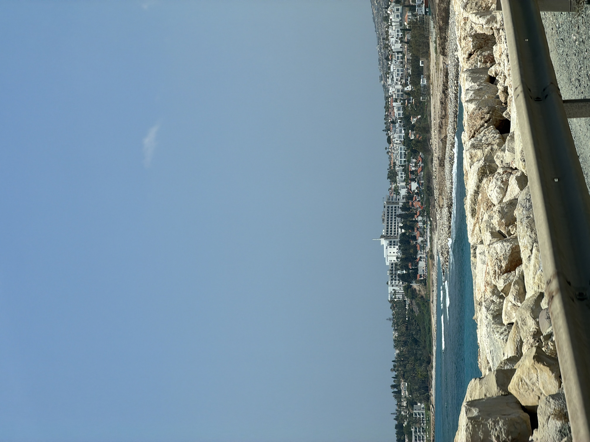 The image shows a coastline with a town in the background. In the foreground, there is a rocky embankment with a metal guardrail. The sea is visible, with waves breaking on the shore. The town consists of white buildings, some of which appear to be hotels or resorts, nestled among trees on a hillside. The sky is clear and blue. There are no people visible in the image.