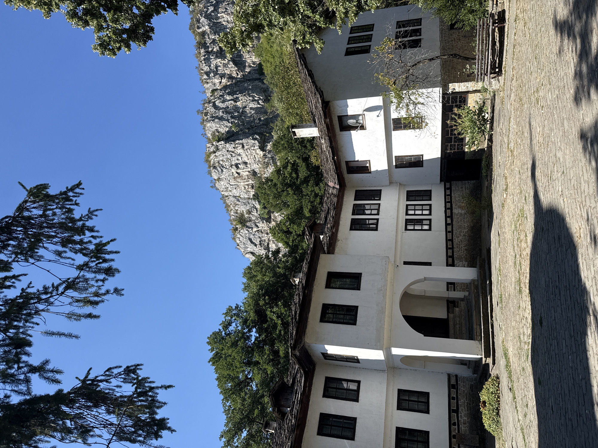 A bright, sunny day with a clear blue sky provides the backdrop for the Dryanovo Monastery in Dryanovo, Bulgaria. The monastery is a two-story white building with dark-framed windows and traditional stone tile roofs. The foreground features a paved courtyard with some greenery and a wooden bench. In the background, lush green trees frame a rocky mountain cliff face. The architecture of the monastery appears historic, with an arched entrance leading to the building's interior. The scene is peaceful and showcases the natural beauty and architectural heritage of the region.