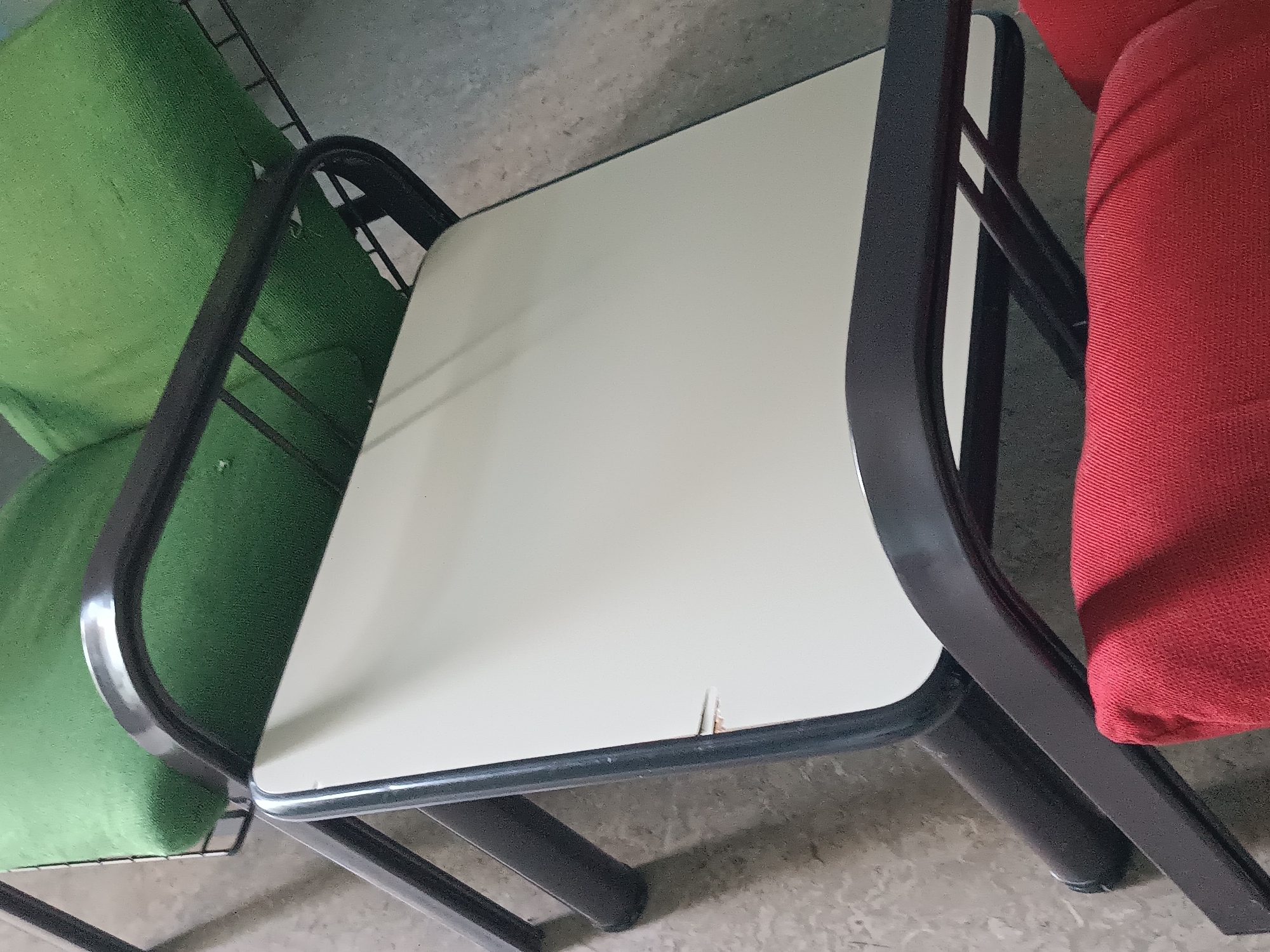 The image shows a close-up view of two chairs, one green and one red, with a cream-colored table in between them. The chairs appear to be made of metal with fabric upholstery. The table has a smooth, flat surface and a dark metal frame. The scene is outdoors, on a paved surface, possibly a patio or a sidewalk. The lighting suggests it is daytime. There are no people visible in the image. The location is identified as Casalmaggiore, Italy.
