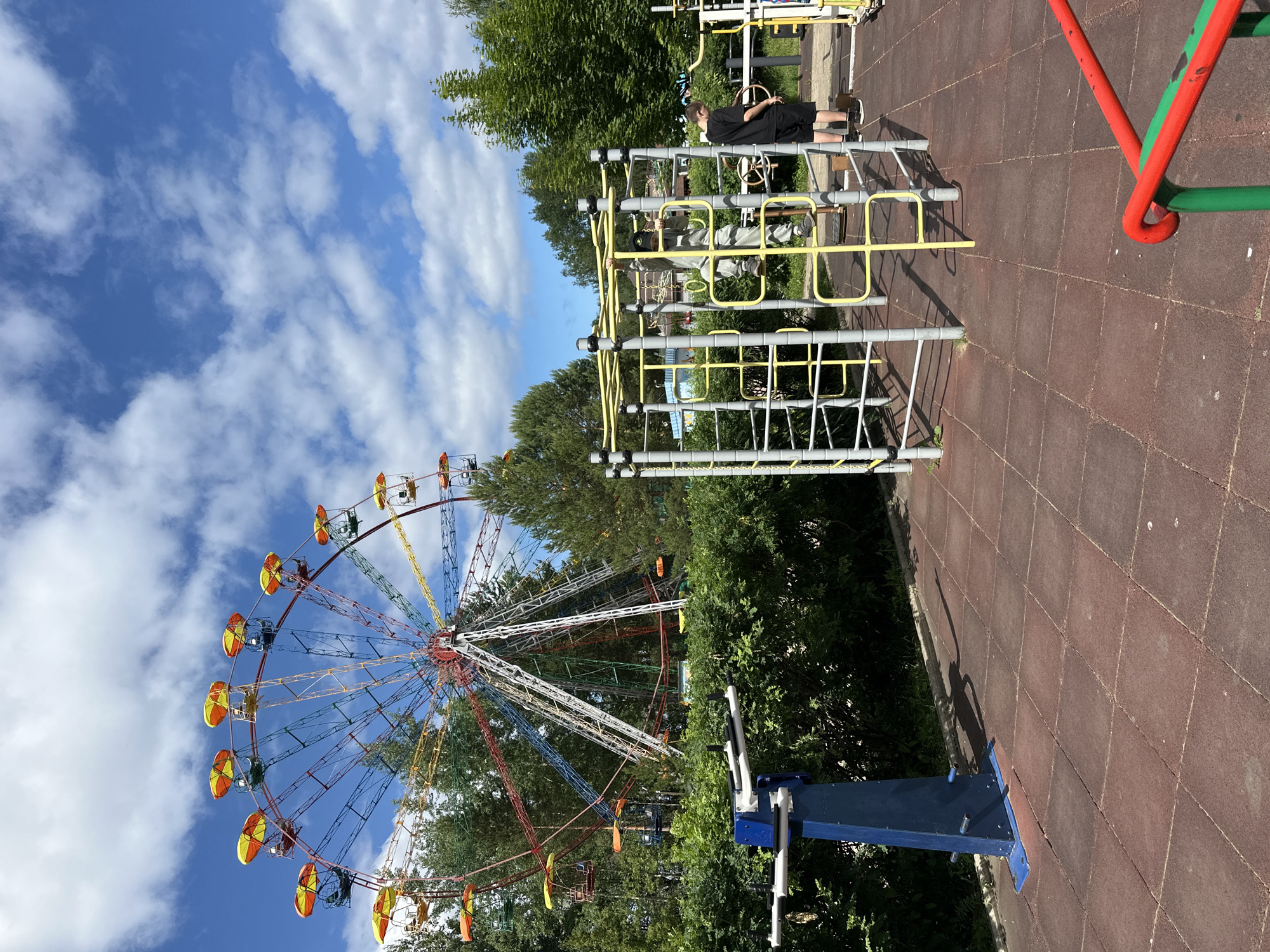 The image depicts an outdoor recreational area, likely in Chaikovsky, Russia, featuring a Ferris wheel in the background and exercise equipment in the foreground. The Ferris wheel is colorful, with red, yellow, blue, and white accents, and is surrounded by green trees under a partly cloudy blue sky. In the middle ground, there is a jungle gym-like structure with yellow and silver bars, and one individual, a male wearing a black t-shirt and shorts, is standing nearby. Another individual, a juvenile wearing grey sweatpants and a hooded jacket, is climbing on the jungle gym. To the left, a blue and white exercise apparatus is visible, set against a backdrop of dense green bushes. The ground surface is composed of red rubberized square tiles. A bright red and green railing is partially visible in the bottom right corner.