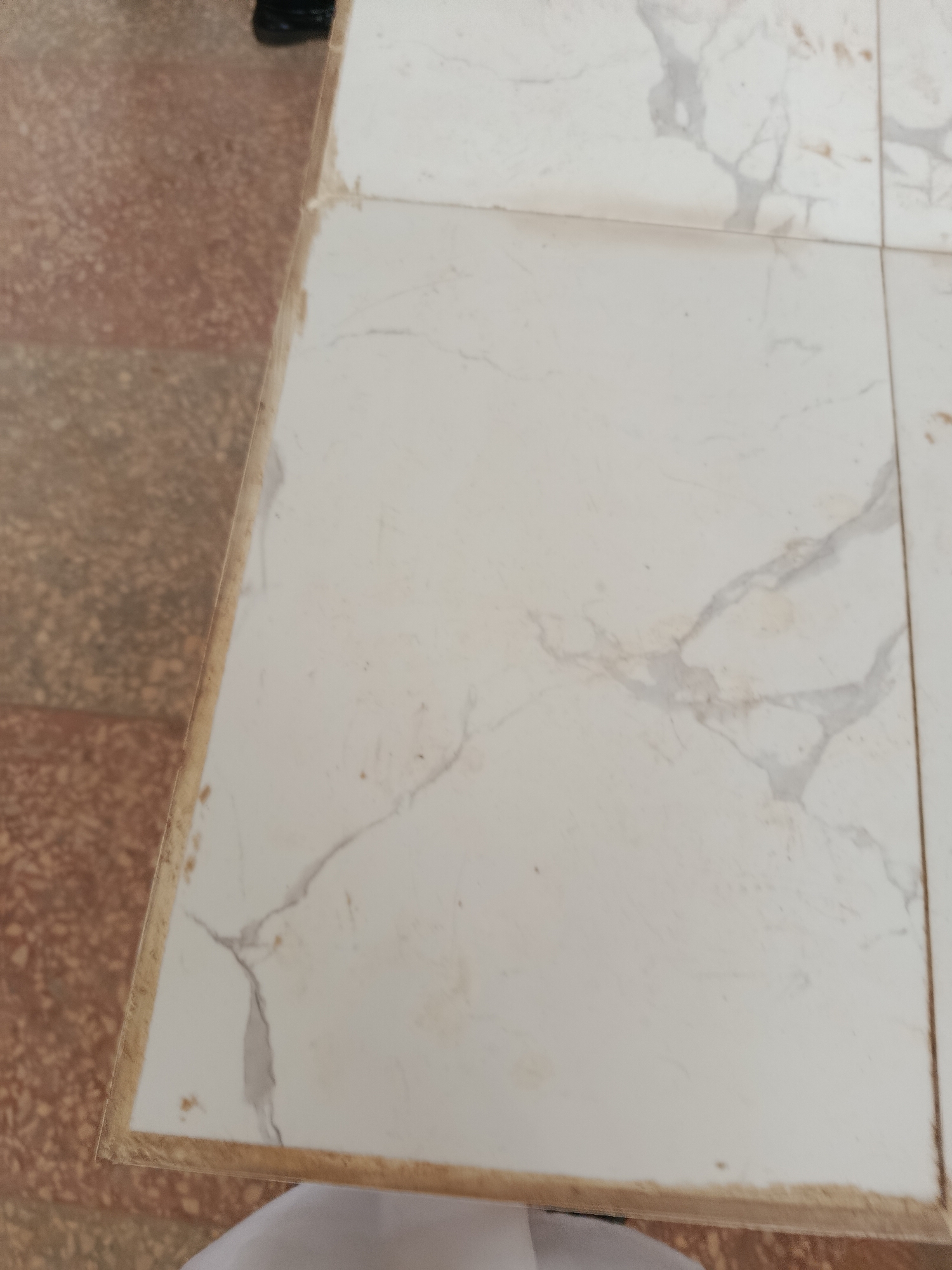 The image shows a close-up of a tiled surface. The tiles are white with grey veining, resembling marble. They appear to be part of a floor or a table. To the left of the white tiles, there is a section of reddish-brown speckled flooring. A thin, light-colored border runs along the edge where the white tiles meet the reddish-brown flooring. The overall impression is of an indoor or outdoor surface. There are no people or specific activities clearly depicted, though the presence of a white fabric at the bottom suggests some human presence or activity. The lighting appears to be neutral, not strongly indicative of a particular time of day. The location context is Ukpor, Nigeria.