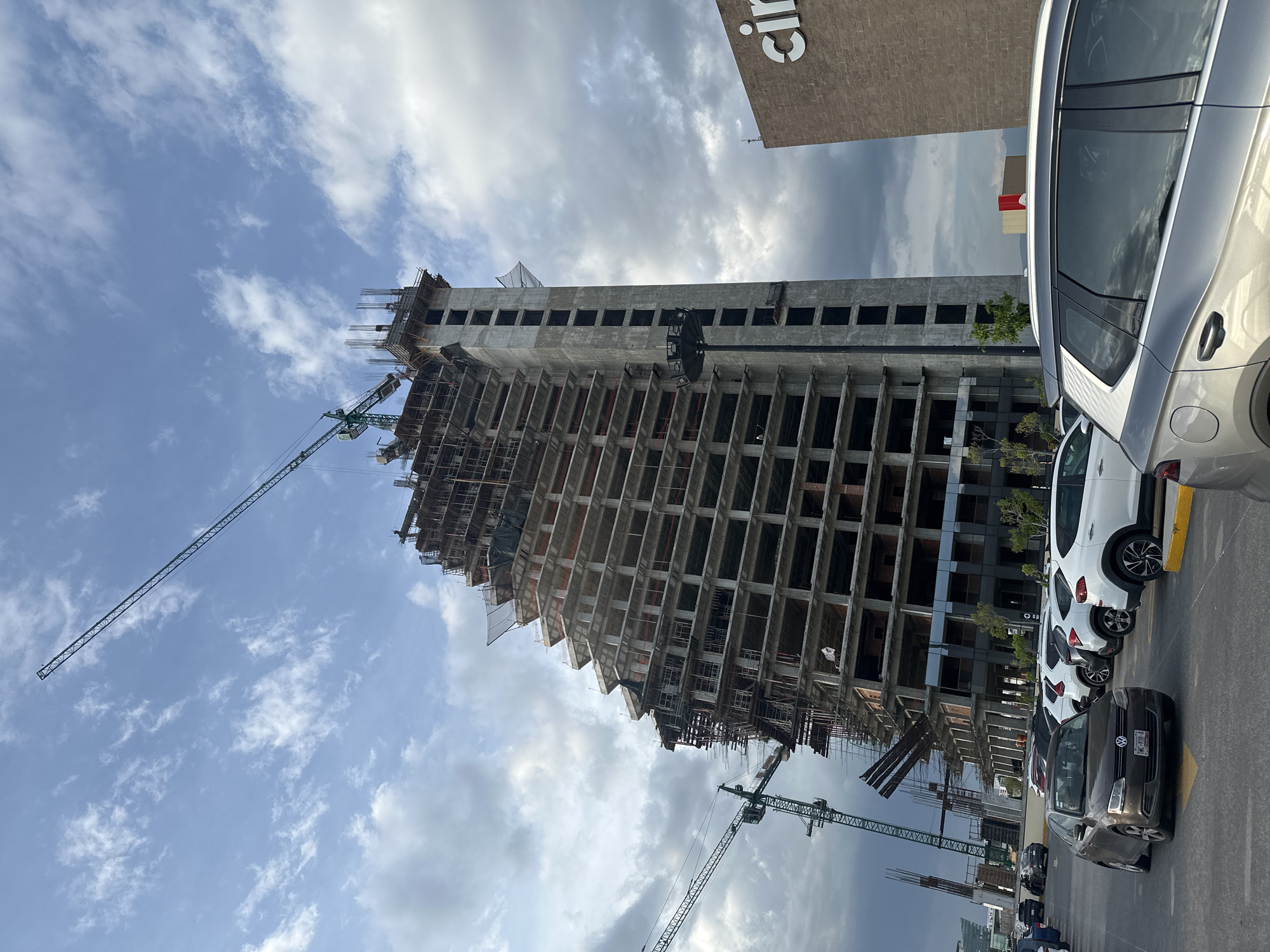 A partially constructed, tall building with a tiered, asymmetrical design dominates the center of the image, appearing to be located in Puebla, Mexico. Two large green construction cranes are visible, one extending diagonally across the sky from the left, and another positioned near the top of the building. The building's exterior reveals exposed concrete floors, rebar, and scaffolding. In the foreground, a parking lot is filled with various cars, including a dark gray sedan, several white SUVs, and a silver SUV. To the right of the construction site, a brick building with a large white "cinemas" logo is partially visible. The sky is partly cloudy, with a mix of bright white clouds and darker, more ominous clouds, suggesting potentially changing weather conditions. The overall scene depicts an active construction zone within an urban environment.