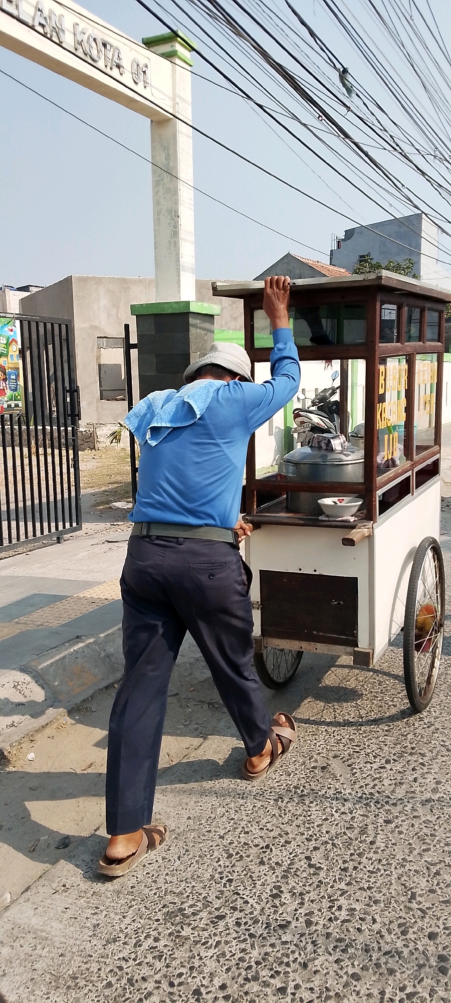 In Babelan, Indonesia, a man wearing a blue long-sleeved shirt, dark pants, and sandals pushes a food cart. A blue towel is draped over his shoulders. The food cart has a wooden frame and glass panels, with what appears to be a large pot inside. The cart is on wheels and is being moved on a paved surface. In the background, there is a metal gate, a building, and a sign that reads "LAN KOTA 01". Overhead, a tangle of electrical wires is visible against a clear sky. The scene suggests a street vendor operating in a developing urban area.