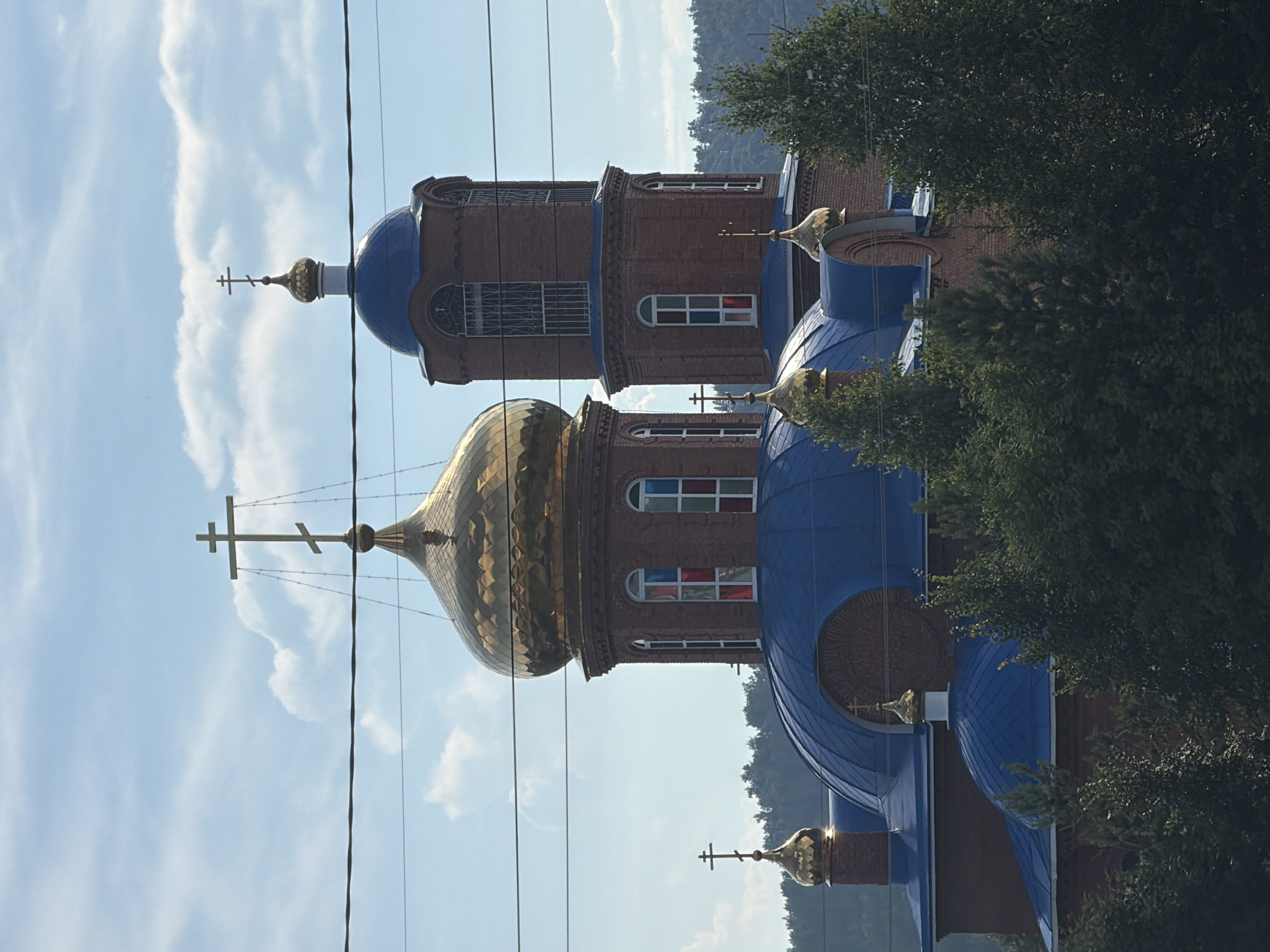 This is an image of a church with blue domes and golden crosses. The church is made of red brick and has several windows. It is surrounded by trees and a hilly landscape. The sky is blue with some clouds, suggesting it is daytime. There are power lines running across the image, partially obscuring the view of the church. The image is taken from a low angle, looking up at the church. The overall impression is one of religious architecture set against a natural backdrop. The image is rotated 90 degrees counterclockwise.