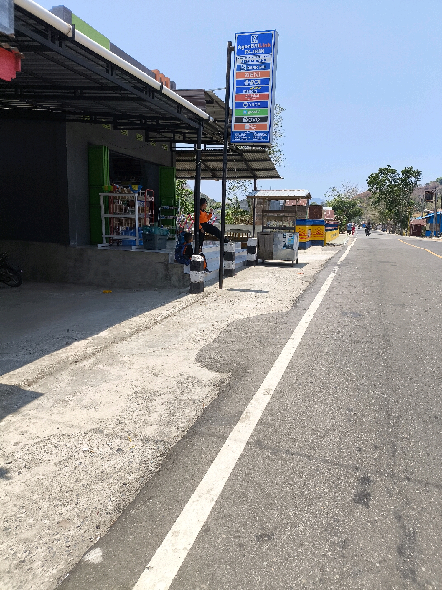 The image depicts an outdoor scene on a sunny day in an urban environment, possibly in Indonesia. A road runs through the center right of the frame, with a white painted line marking its edge. To the left of the road is a concrete sidewalk and a small shop. The shop has a dark gray facade and a green folding door. A black metal awning extends over the sidewalk in front of the shop.
A tall blue sign, labeled "AgenBRILink FAJRIN", stands near the shop. The sign lists various financial services and brands, including "SEMUA BANK", "BANK BRI", "BNI", "BCA", "mandiri LinkAja!", "DANA", "GOPAY", "OVO", and "DANA". Below these logos, it details services such as "BAYAR LISTRIK" and "BAYAR PDAM".
Two individuals are visible near the shop. One, wearing an orange shirt, is seated on a low wall to the right of the shop. Another, a child with a blue backpack, is sitting on the stairs leading up to the shop's entrance. A motorcycle is parked on the left side of the frame, near the shop.
In the background, the road continues into the distance, with a few buildings and trees lining its sides. The landscape suggests a hilly or mountainous terrain in the far background, under a clear blue sky. The scene appears to be a typical street view of a small town or a commercial area in Indonesia.