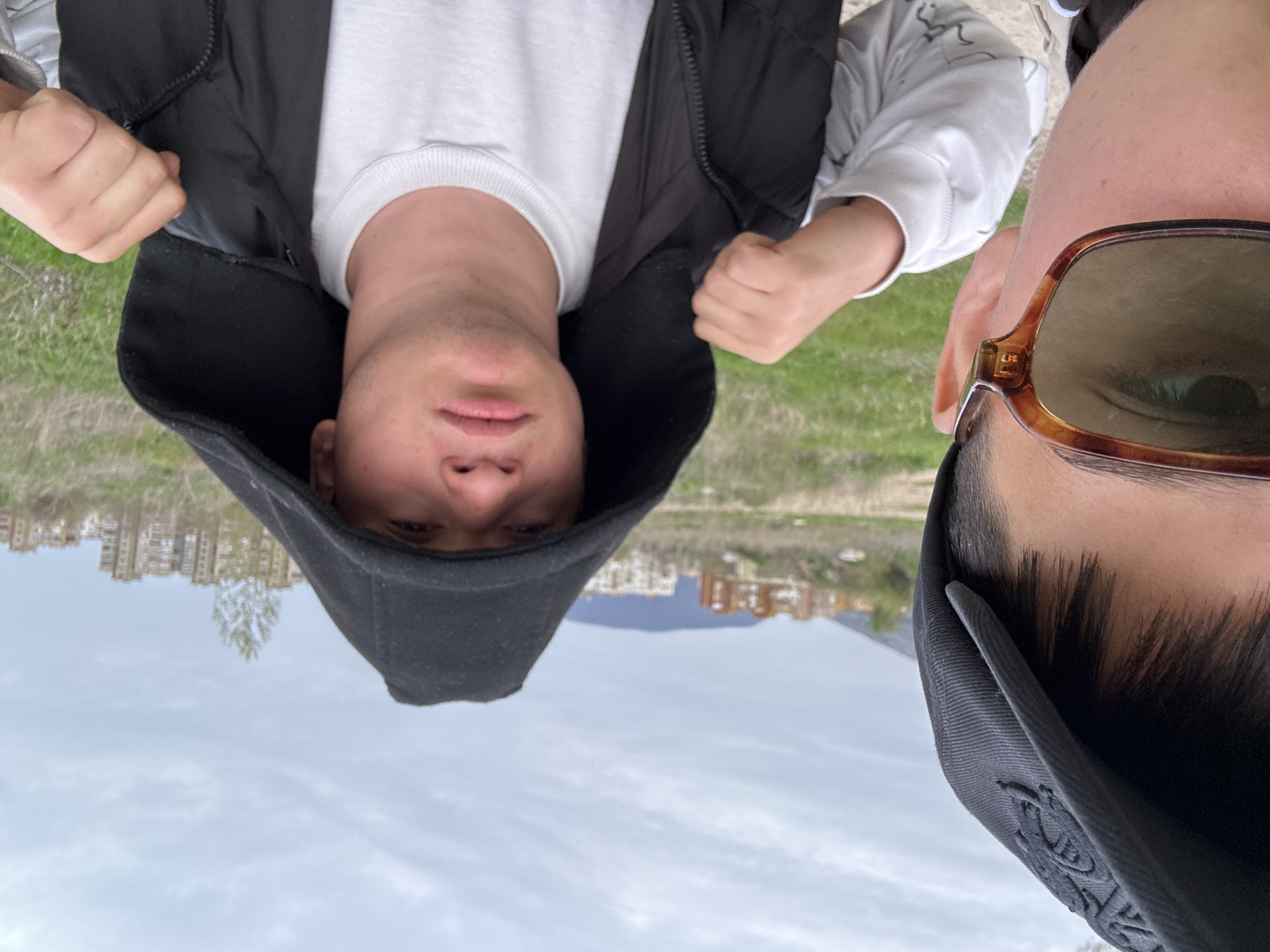 The image shows two people, one wearing a black hooded jacket and a white sweater, and the other wearing sunglasses and a black cap. Both are positioned close to the camera, creating a selfie-style composition. The background features buildings and a distant mountain range under a cloudy sky. The photo appears to have been taken in the city of Unknown, Unknown.