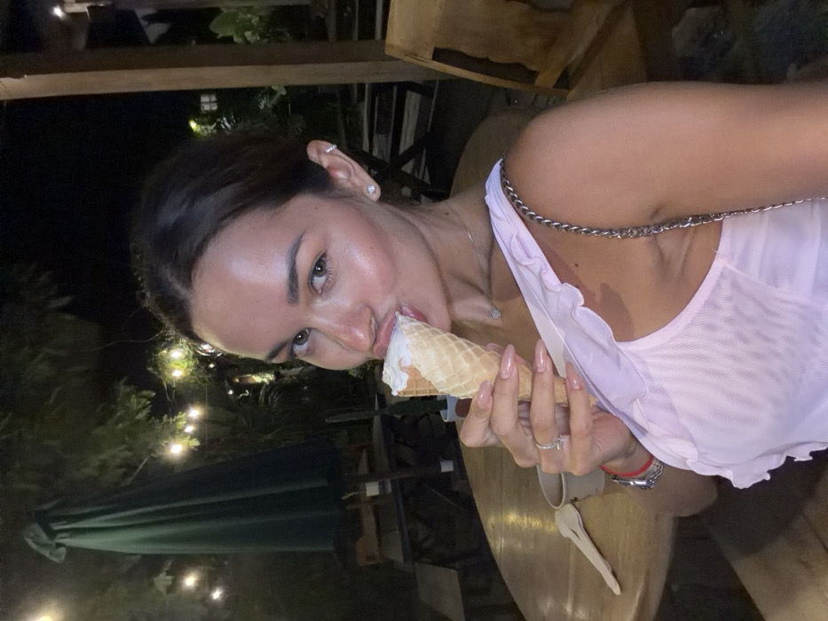 An outdoor, nighttime scene in Pecatu, Indonesia, depicts a female individual with dark hair and a light complexion, photographed from the chest up. She wears a light pink, possibly sheer, ruffled top and a silver chain strap from a bag is visible on her right shoulder. She is actively eating from a sugar cone filled with white ice cream, held in her left hand, with her mouth partially around the ice cream. Her left hand displays a silver ring on the fourth finger and a red bracelet on the wrist. She has stud earrings in her earlobes and a cuff earring on her left helix. The background is dimly lit by string lights hanging amidst dark foliage and trees. A round wooden table is visible, on which a light-colored cup and a wooden spoon are present to the left of the subject. A large, dark green outdoor umbrella is partially visible behind her to the left, and dark wooden outdoor furniture is discernible further back.