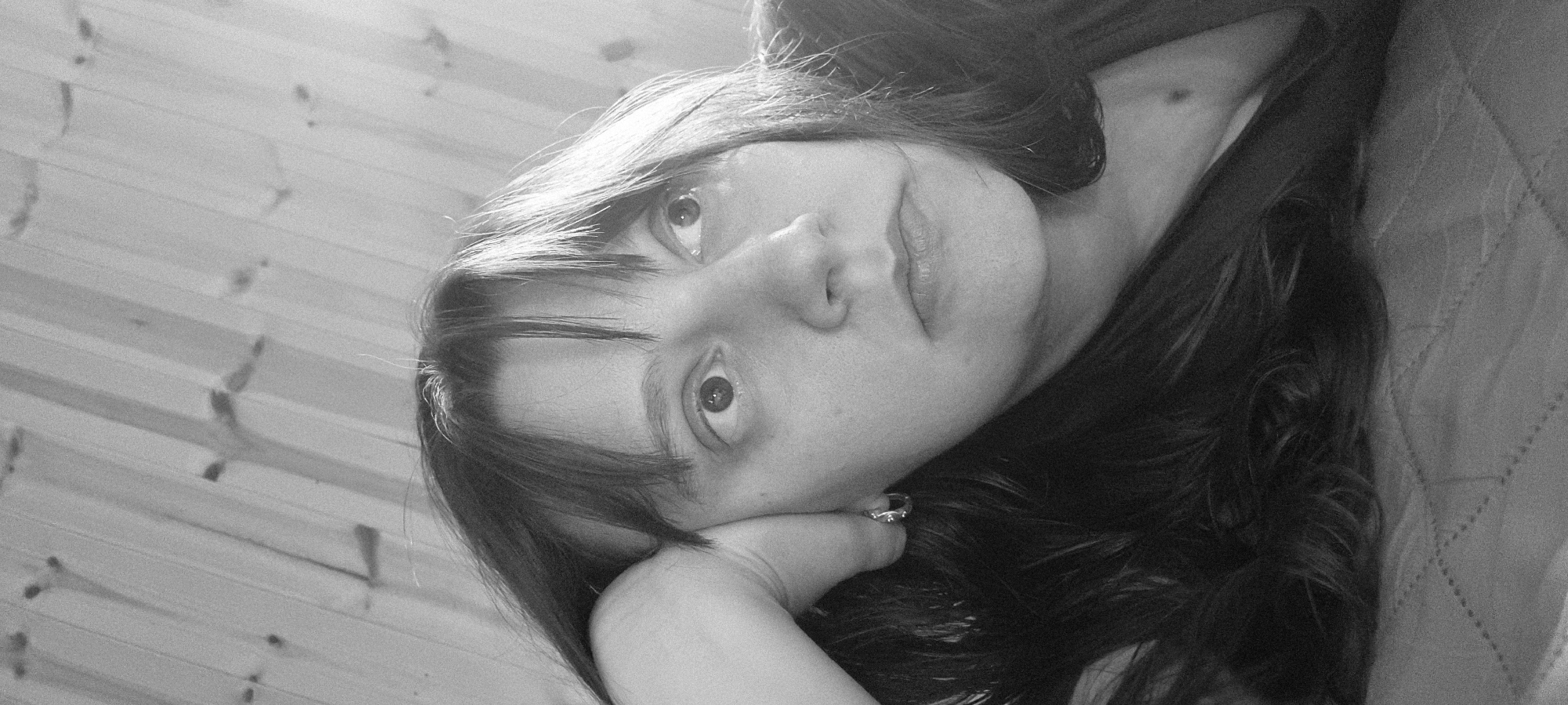 A monochrome, close-up portrait depicts a young woman with dark hair and fair skin. She is reclining, her face angled upwards and slightly tilted to the side, resting on her hand. She is wearing a ring on her finger. The background includes a wooden wall and a quilted surface. The location is not specified.