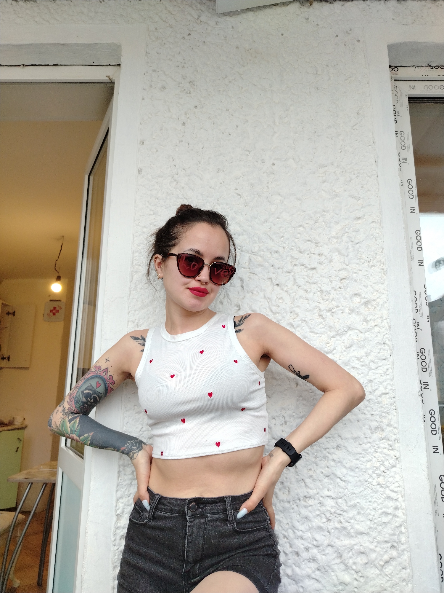 A woman stands between two white window frames against a white wall. She is wearing a white crop top with red hearts, black shorts, and red-tinted sunglasses. Her hair is pulled back in a bun. She has tattoos on both arms. She rests her hands on her hips. Through the open window to her left, part of a room with light-colored walls, a light fixture, a green cupboard, and a table with a stool are visible. The letters "GOOD IN" are vertically printed several times on the window frame to her right. The city of Noviy, Russia, is not discernible from the image.