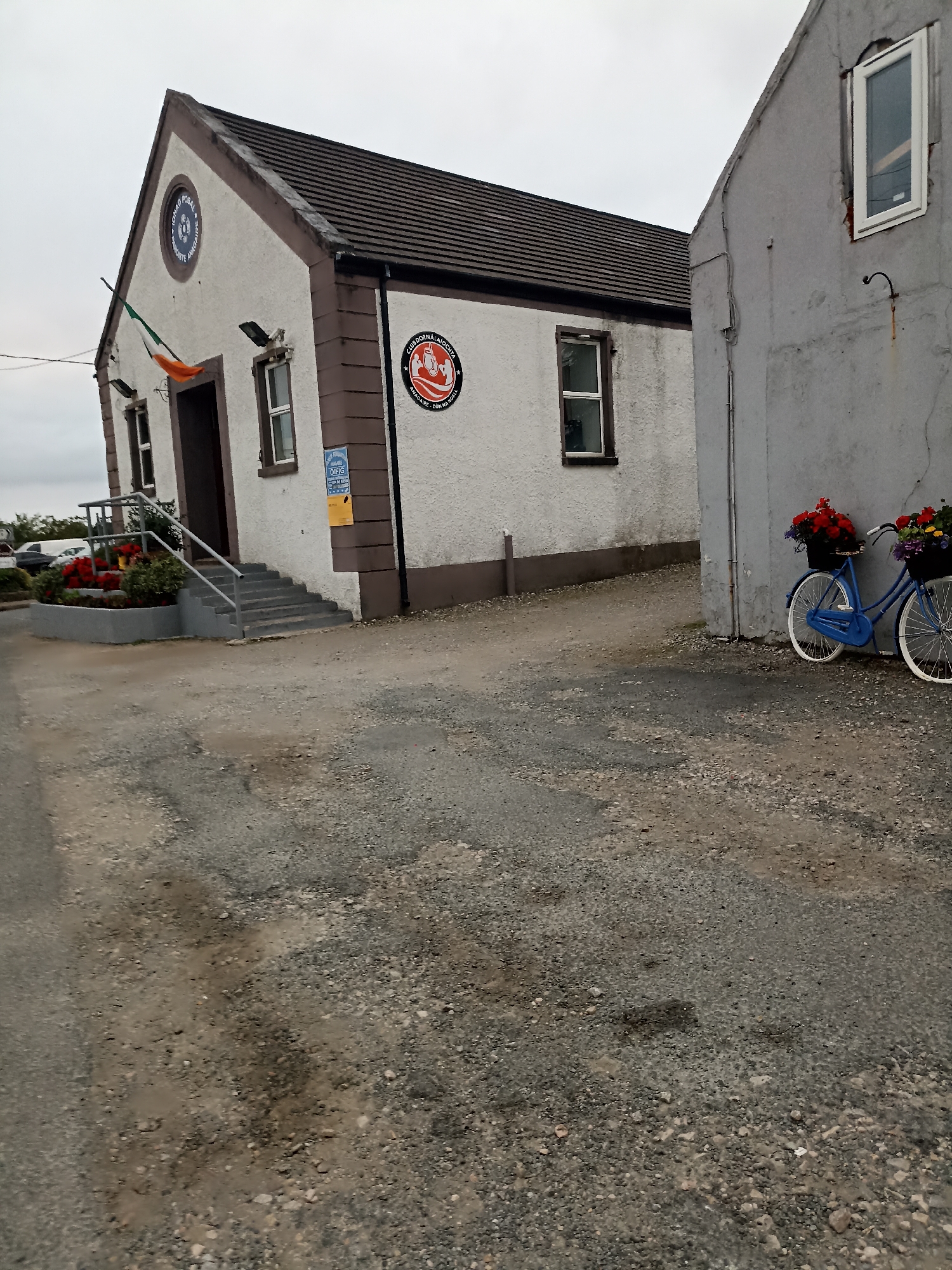 The image shows the exterior of a building, likely a community hall or club, in an outdoor setting. The building is white with a brown trim and a dark, sloped roof. Two circular emblems are visible on the front of the building, one with a green, white, and orange flag draped below it. A set of grey stairs leads up to the entrance. To the right of the building, another structure, partially visible, is a light grey color. A blue bicycle adorned with flower baskets is leaning against this grey structure. The ground in front of the buildings is a mixture of gravel and dirt. The sky is overcast. The scene appears to be in a rural or suburban area of Ireland, given the flag and the style of the buildings. There are no people visible in the image.