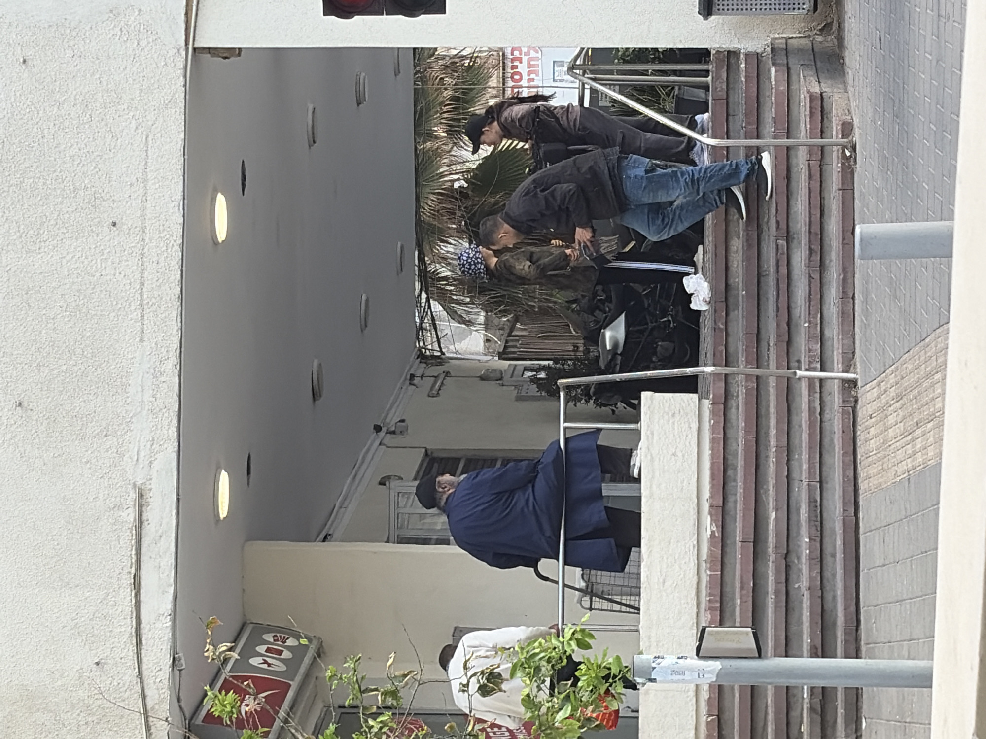 The image depicts a street scene in Tel Aviv, Israel. Several individuals are visible: Three people are walking up the staircase with handrails, another person is leaning out from the window and another person is standing below. The architectural setting includes buildings with light-colored walls, windows, and what appears to be a stairway leading up to a higher level. There is a storefront with a red and white sign, and some greenery. A utility pole with stickers on it is also present. The time of day appears to be daytime, with good lighting.