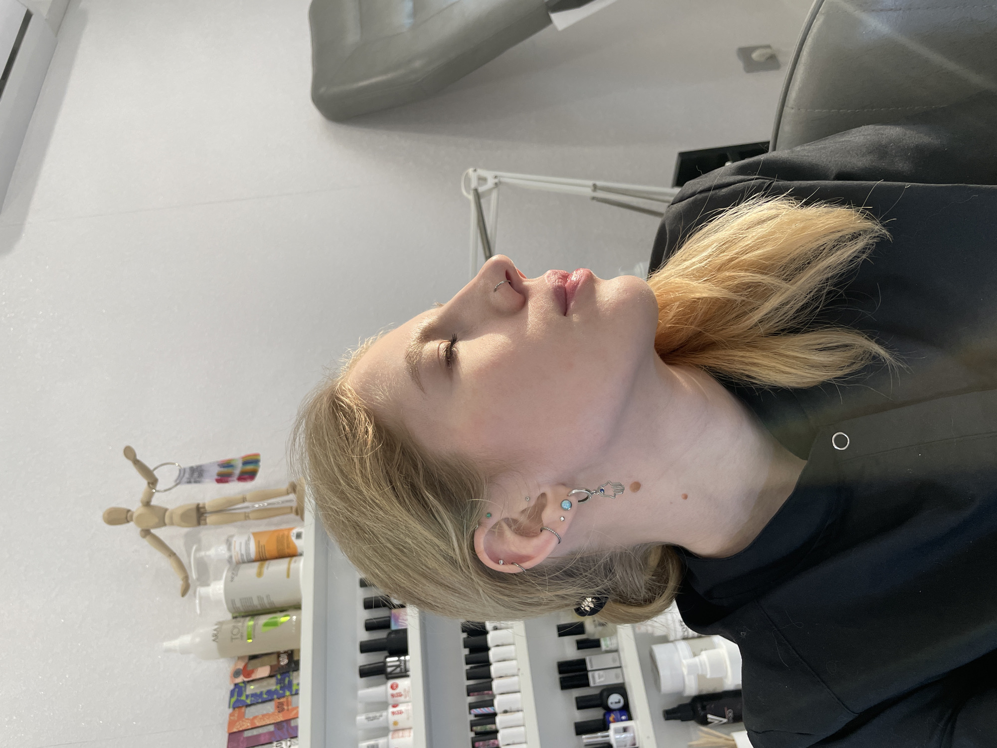 A medium shot shows a fair-skinned woman with blond hair, who is tilted back with her head to the left. Her eyes are closed and she has a silver ring in her left nostril. A silver Hamsa hand earring is visible in her left ear. A black garment with a white button at the neckline is wrapped around her shoulders. Behind her is a gray chair and to the left, a white shelving unit contains rows of cosmetic products and other items. At the top of the shelving unit is a small wooden mannequin. The location and city are not determined in the image.