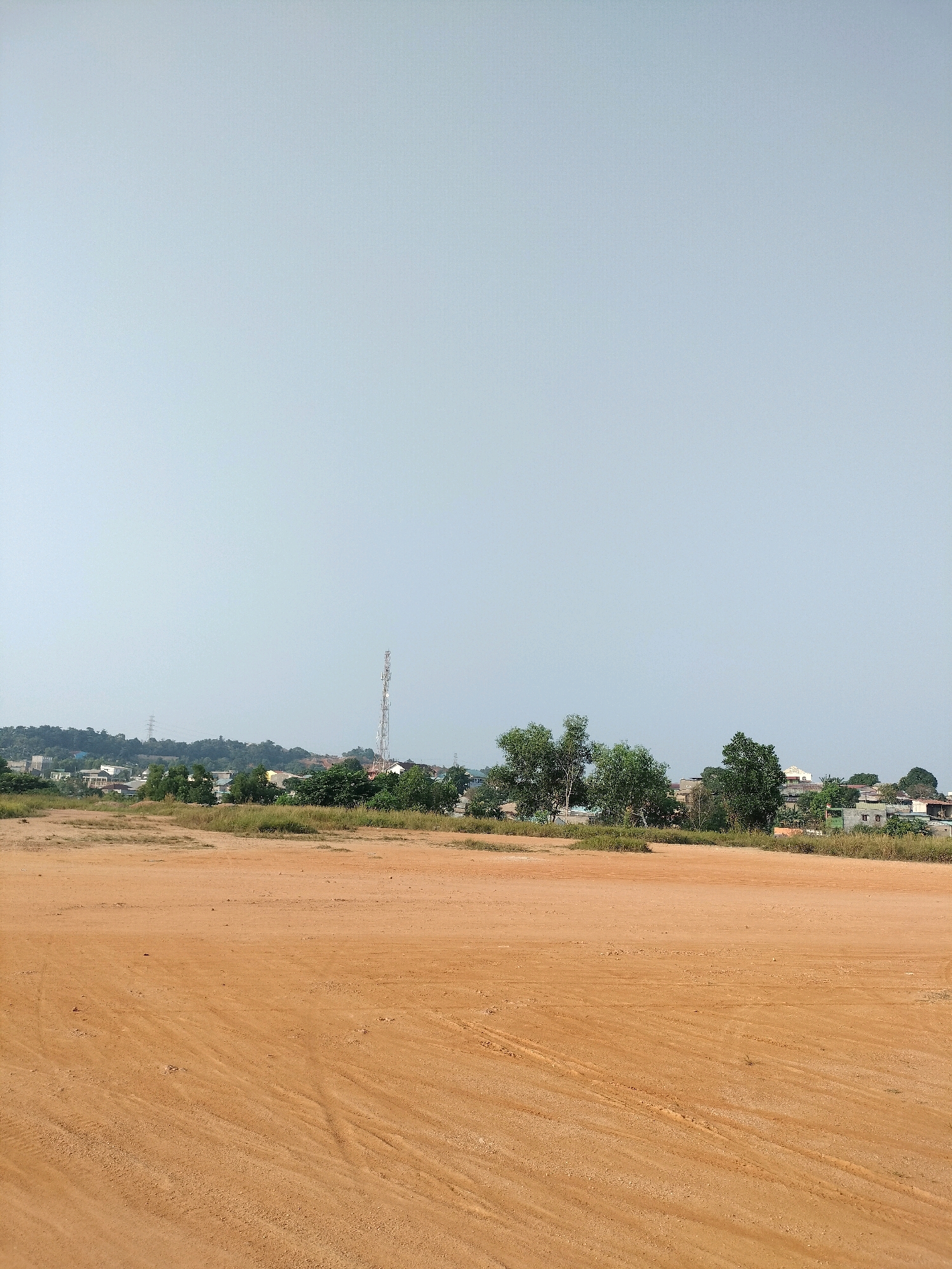 The image captures an open, dusty field of orange soil in the foreground. The ground is uneven with visible tire tracks and scattered small debris. In the middle ground, a line of green vegetation and trees separates the field from a cluster of buildings. A tall, silver telecommunications tower stands prominently among these buildings, which appear to be residential or commercial structures with various roof colors. Further in the background, rolling hills are covered in dense green foliage. The sky is a pale, hazy blue, suggesting a clear but possibly humid day. The scene is likely a developing or semi-urban area within Batam, Indonesia, given the presence of both natural landscape and infrastructure typical of such locations. There are no people or animals visible in the frame.