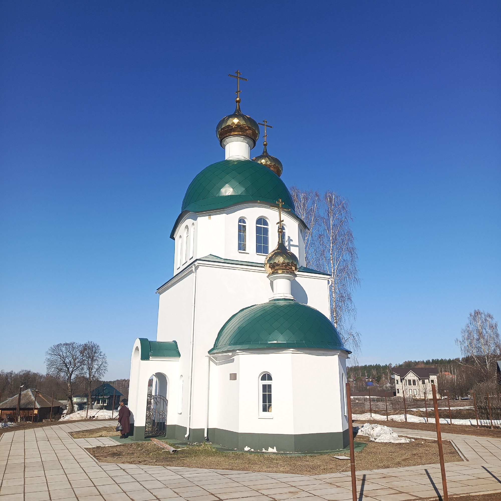 The image shows a white Orthodox church with green roofs and golden domes in Markovo, Russia. The church has a main structure and a smaller, rounded section attached to it. Each section has a green roof and a golden dome topped with a cross. The building has arched windows. A paved walkway leads to the church's entrance, and a person stands near the entrance. The background features trees, other buildings, and a clear blue sky. Snow patches are visible on the ground.