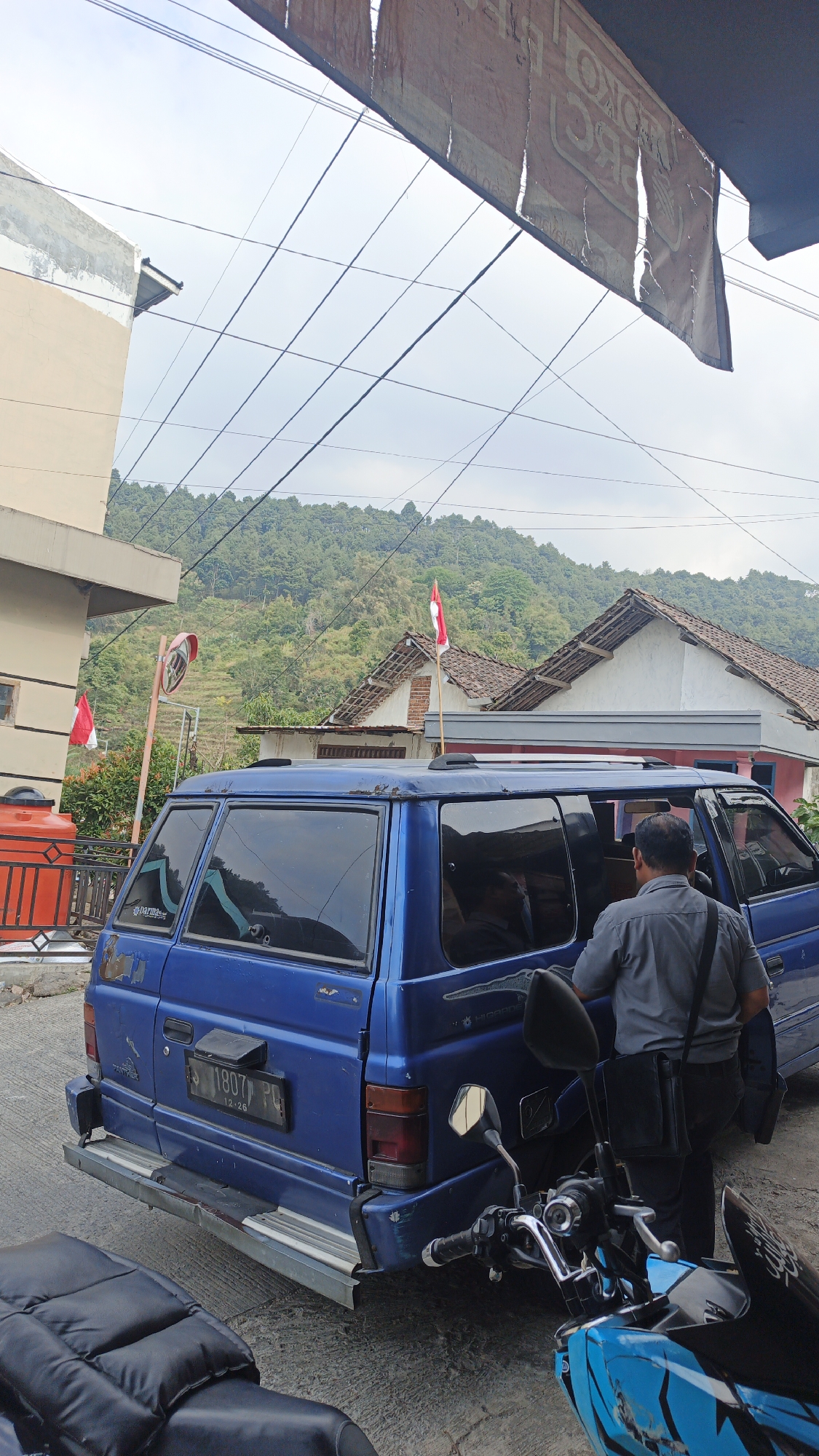 A medium-shot, eye-level photograph captures a street scene in Sajen, Indonesia, under an overcast sky. In the foreground, a motorcycle with a blue and black patterned fairing is parked on the right. To the left, a portion of another motorcycle's black seat is visible. Dominating the mid-ground is the rear and side of a blue minivan. A man in a gray short-sleeved shirt and dark pants is standing by the open passenger door of the minivan, facing away from the camera, with a black bag slung over his shoulder. The minivan has a license plate reading "1807 PG" with "12-26" below it. Behind the minivan, a traditional Indonesian house with a brown tiled roof is partially visible. Further in the background, a lush, green hillside is terraced with vegetation, indicating agricultural activity. Two Indonesian flags, one on a tall pole near the house and another on a shorter pole to the left, are prominently displayed. To the far left, a light-colored building with a white and beige facade stands, with an orange water tank visible on its left side. Overhead, a network of electrical wires crisscrosses the sky, and a tattered, brown banner hangs from above, partially obscuring the view. The overall impression is of a typical, lived-in neighborhood scene in rural Indonesia.