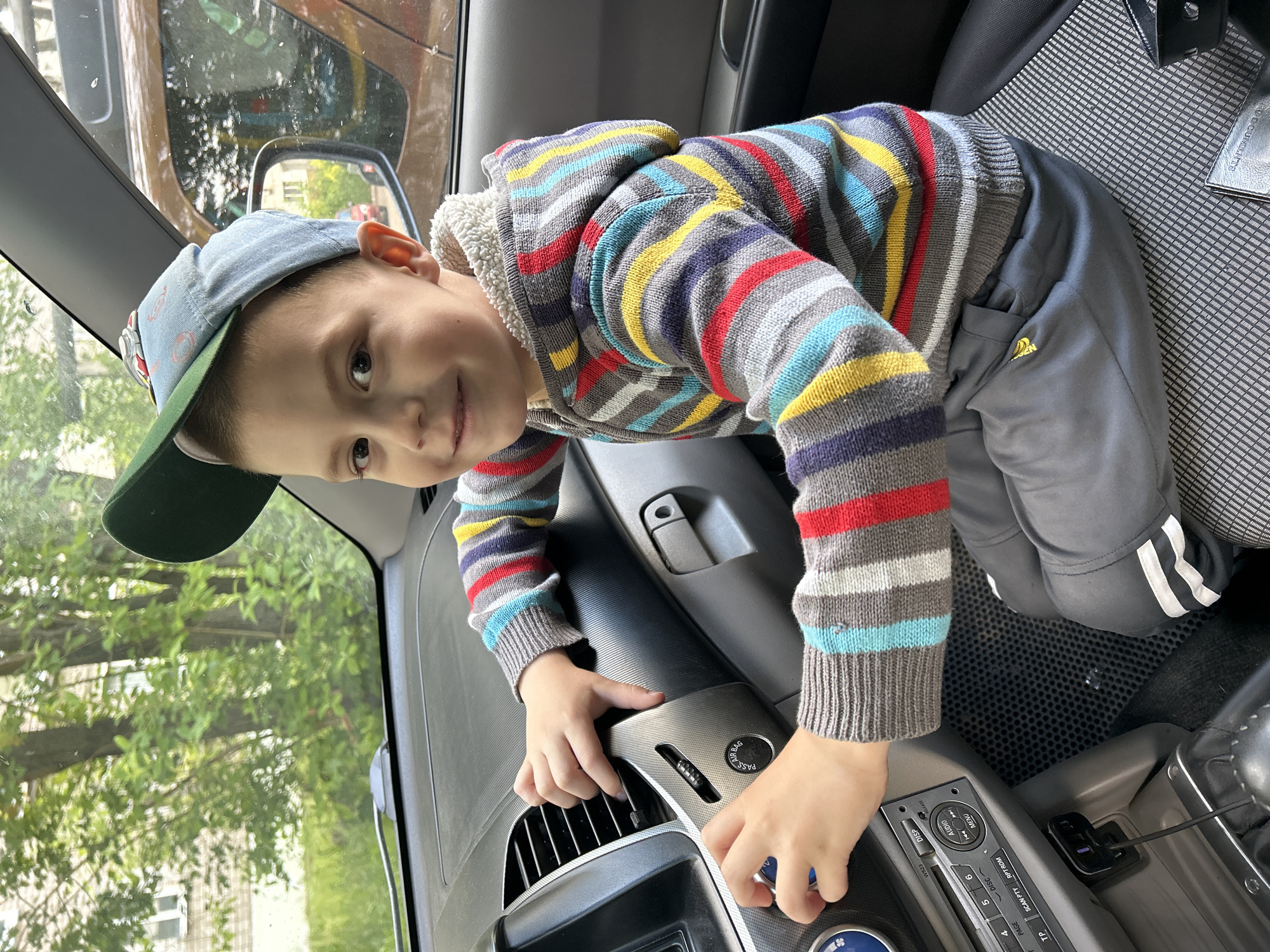 A young boy wearing a striped hoodie and a baseball cap sits in the driver's seat of a car. He is looking directly at the camera with a slight smile. His hands are on the car's dashboard, appearing to interact with the controls. The interior of the car is visible, including the dashboard, air vents, and a car stereo. Outside the car, trees and a building can be seen through the windows. The location is the city of Chaikovsky, Russia.