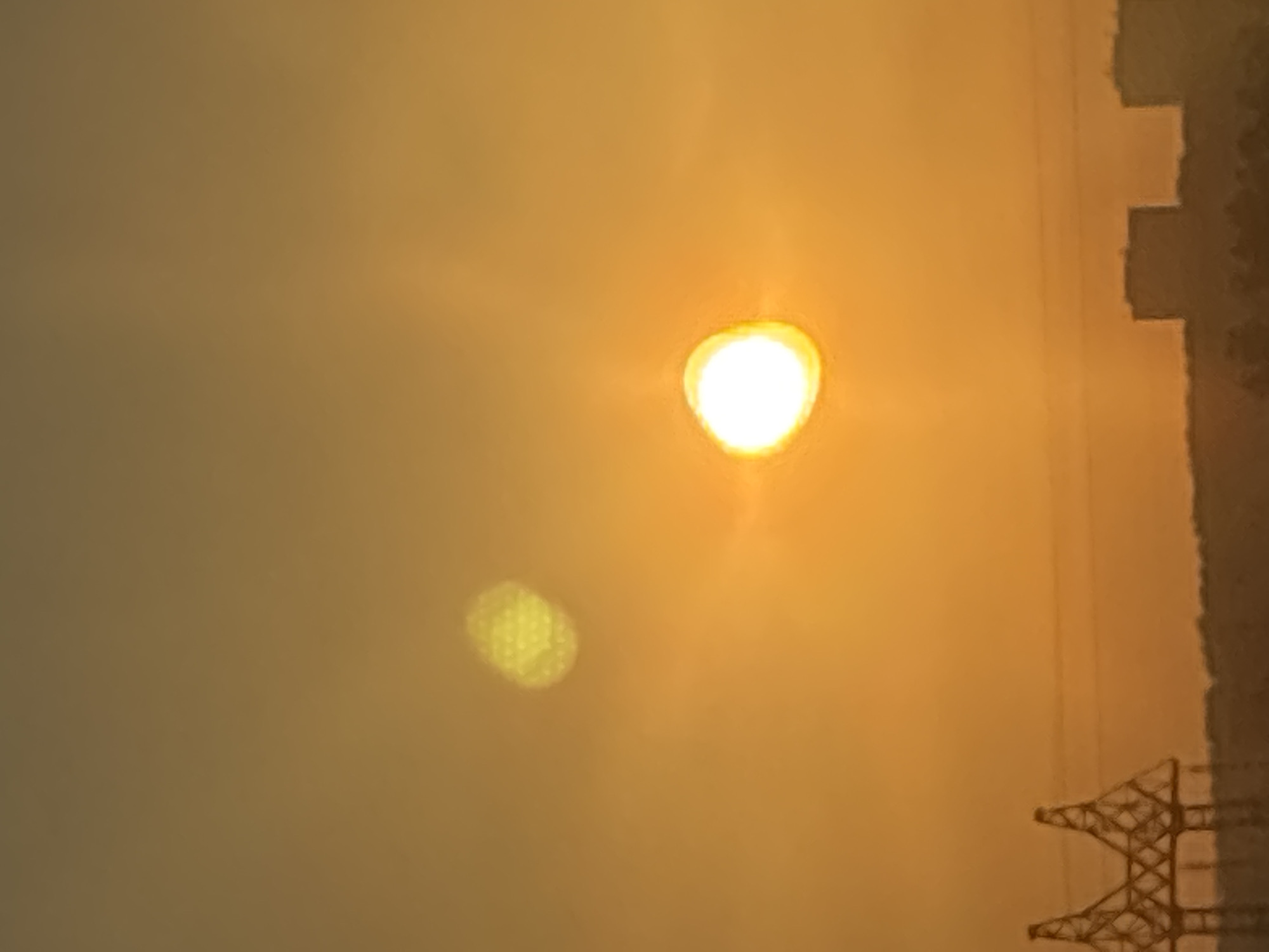 The image presents an outdoor scene over the city of Chaikovsky, Russia, at either sunrise or sunset, characterized by an orange-yellow sky. A bright, white-yellow sun, displaying visible lens flare, is prominently positioned in the lower-central portion of the frame. Above and slightly to the left of the sun, a distinct, soft, yellow-green lens flare artifact is also present. Along the distant horizon, a dark silhouette reveals structures and terrain. On the left side, a large, dark, lattice-patterned structure, consistent with a power transmission tower or a section of a large bridge like those found near the Chaikovsky Hydroelectric Station, is partially visible. To the right, several distant, dark, rectangular building silhouettes rise above a subtle, undulating dark line suggesting trees or distant land. No individuals or specific activities are discernible.