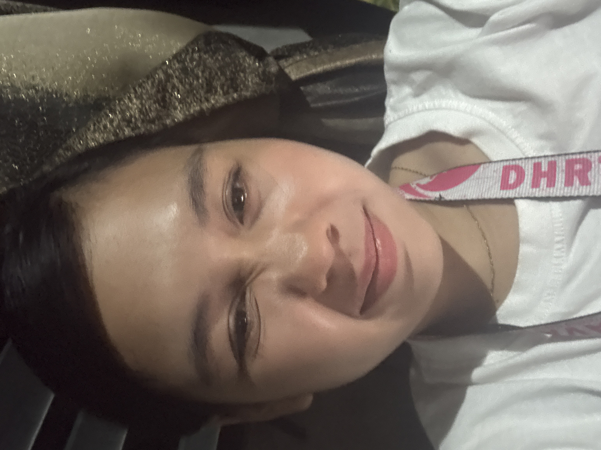 The image is a selfie of a young woman in Davao City, Philippines. She has dark hair pulled back and is wearing a white t-shirt. A lanyard with the letters "DHR" in pink is visible around her neck. She is making a face, squinting her eyes and pursing her lips. The background appears to be indoors, with blinds visible on the left and a textured fabric in a gold and dark brown color on the right. The lighting is dim, creating highlights on her face.