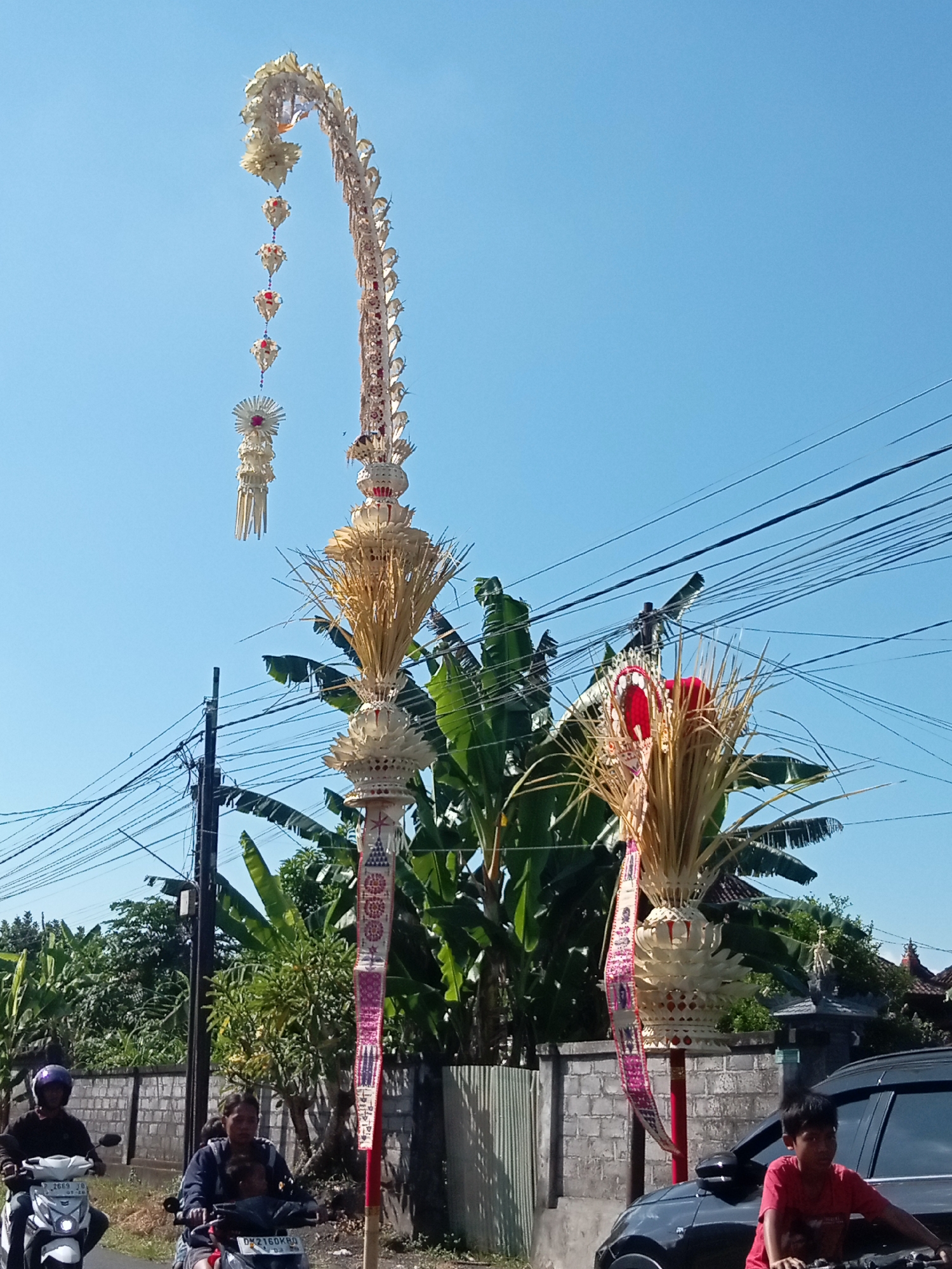 In an outdoor scene, two tall, ornate, arched structures decorated with woven palm leaves and red and white ribbons dominate the foreground under a clear blue sky. The structure on the left curves upward and is adorned with several dangling, decorative elements. The structure on the right, shorter and straighter, also features woven palm leaves and ribbons. Several power lines are visible crisscrossing the upper portion of the image.
In the middle ground, lush green banana trees and other vegetation are present, partially obscuring a grey brick wall. A utility pole stands to the left of the taller structure.
In the foreground, on a paved road, two individuals are riding motorcycles. The person on the left, wearing a blue shirt and purple helmet, rides a white motorcycle. The person to their right, wearing a dark jacket, rides a motorcycle with a visible license plate. A black car is parked on the right side of the road, with a young person in a red shirt leaning against it. The setting appears to be a street in a tropical or subtropical region.