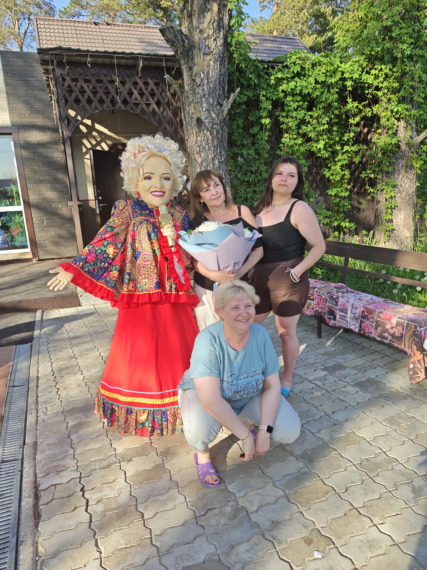 The image shows four individuals in what appears to be a park or garden setting. On the left stands a life-size doll dressed in a red and floral patterned traditional Russian dress holding a microphone. Behind the doll are two women. The woman on the left has medium-length hair and is holding a bouquet of flowers wrapped in light blue paper. The woman on the right is younger, wearing a black top and brown shorts. In front of them, squatting on the paved surface, is a woman with short blonde hair, wearing a light blue T-shirt and grey pants, with purple sandals.
The setting features a building with dark wooden latticework, and greenery visible in the background, as well as a tree. A bench covered with a patterned cloth sits to the right. The pavement has a distinct tessellated pattern.
The city is Chaikovsky, Russia.