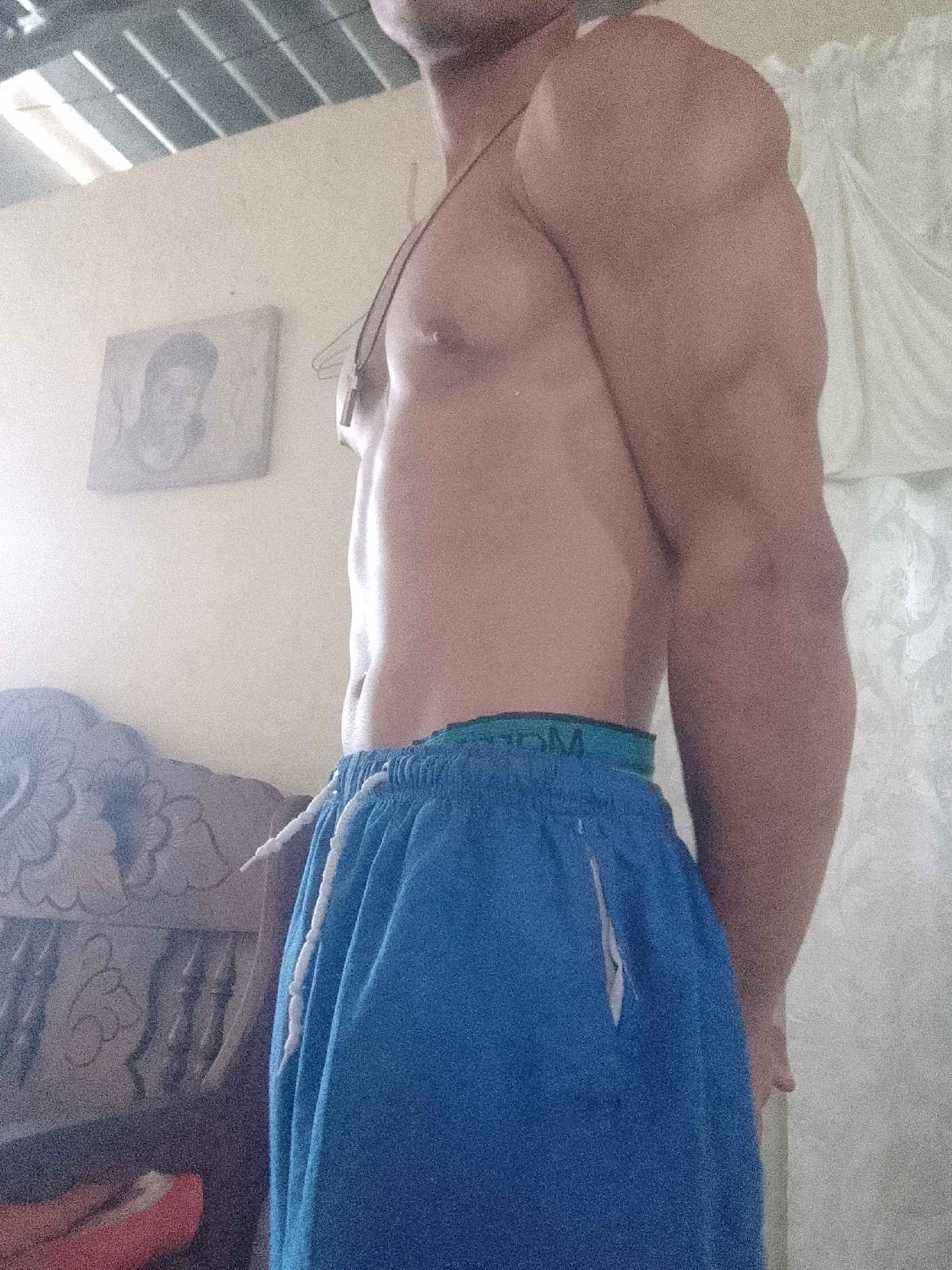 A shirtless man is standing indoors wearing blue drawstring shorts. He has well-defined muscles. A framed picture hangs on the wall behind him to his left, and a carved wooden chair is visible. White curtains are on the right. The location is not identifiable as Juan Griego, Venezuela, based on the given information.