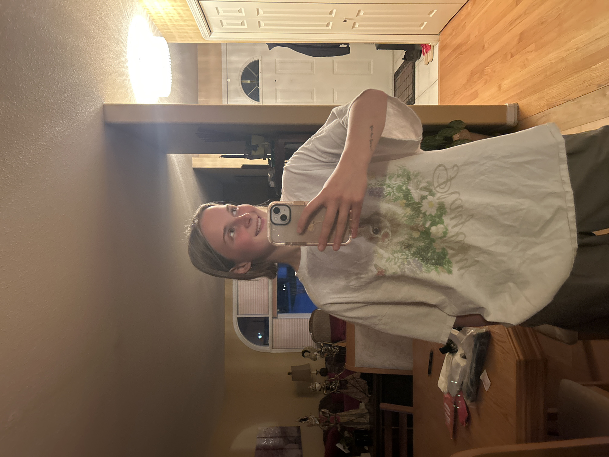 A young, fair-skinned person with short blonde hair is holding a smartphone to take a selfie in what appears to be the interior of a home. The person is wearing a white t-shirt with a floral design and what looks like grey pants. The background suggests an interior hallway or entrance area with a front door visible. There's a chandelier-type light fixture above, and a wooden floor can be seen leading away from the entrance. In the reflection, a dining area with furniture and decorative items can be observed. Determining the specific location within Kelowna, Canada from the image alone is not possible without additional information or context.