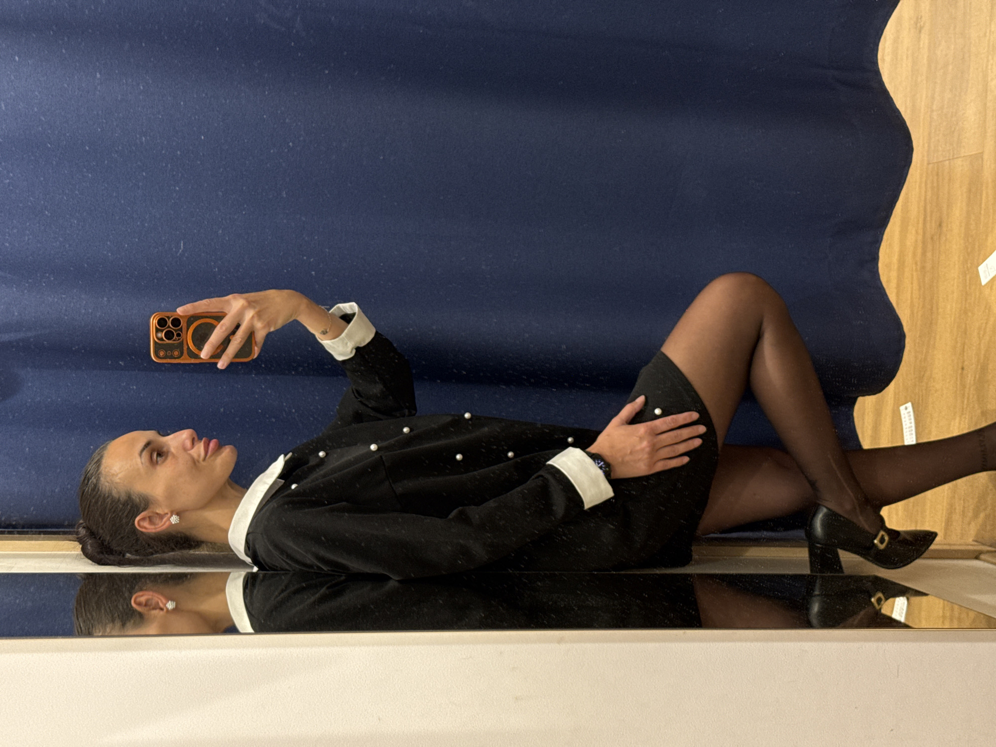 A woman is lying down, taking a selfie with an orange phone. She is wearing a black dress with white collar and cuffs, black stockings, and black high heels. She is lying on a mirrored surface. The background is a blue and brown wall. The image does not contain enough information to provide the location in Pleven, Bulgaria.