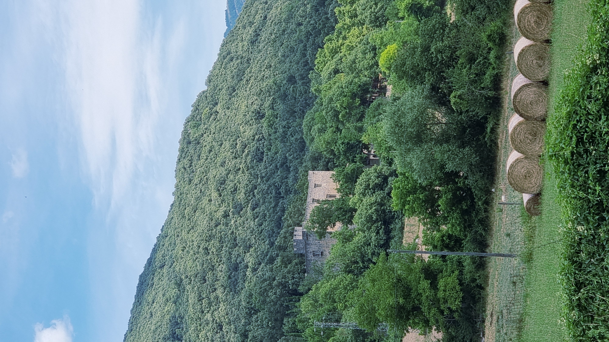 In Montagut i Oix, Spain, a stone castle stands nestled amongst lush green trees on a hillside. In the foreground, several round bales of hay lie in a grassy field under a partly cloudy blue sky. A solitary utility pole is visible to the left of the castle, and a faint fence runs through the field. The castle appears to be an old, possibly historic structure with multiple windows and battlements. The overall scene depicts a rural landscape with a touch of history.