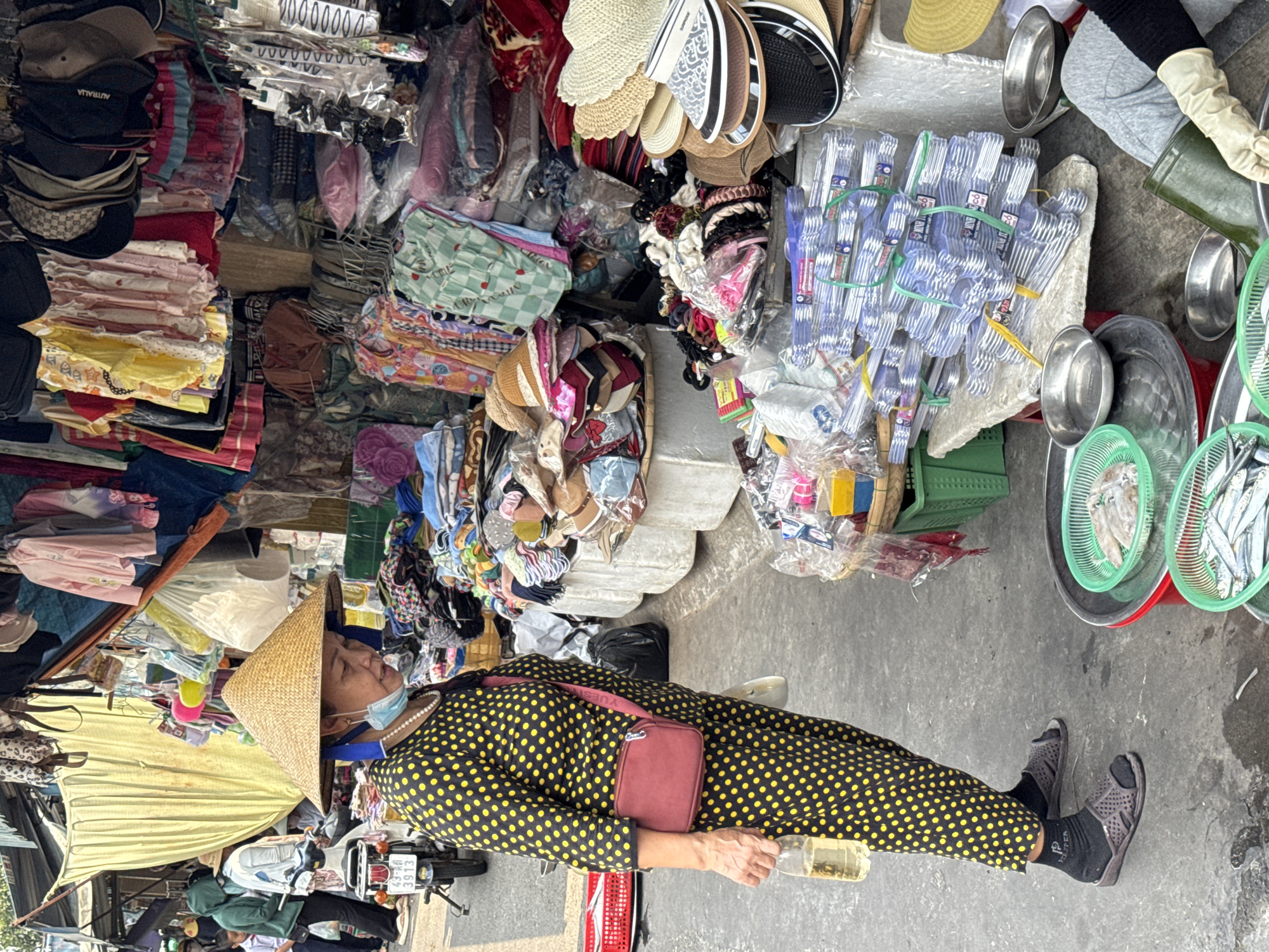 A high-angle, full-frame outdoor shot depicts an Asian woman wearing a conical straw hat, a black and yellow polka dot dress, a strand of pearls, black socks, and open sandals. She also has a red cross-body bag. She is standing with a glass bottle in her hand on a concrete surface.
The woman is positioned near a cluttered market stall or shop, filled with various goods. These include hats, clothing, and hanging items. To her right, a metal tray holds small fish. The environment suggests an outdoor market in a city in Vietnam. The woman's appearance indicates she is possibly a vendor or local resident. The photo captures a moment in the bustling environment of a market in Vietnam.