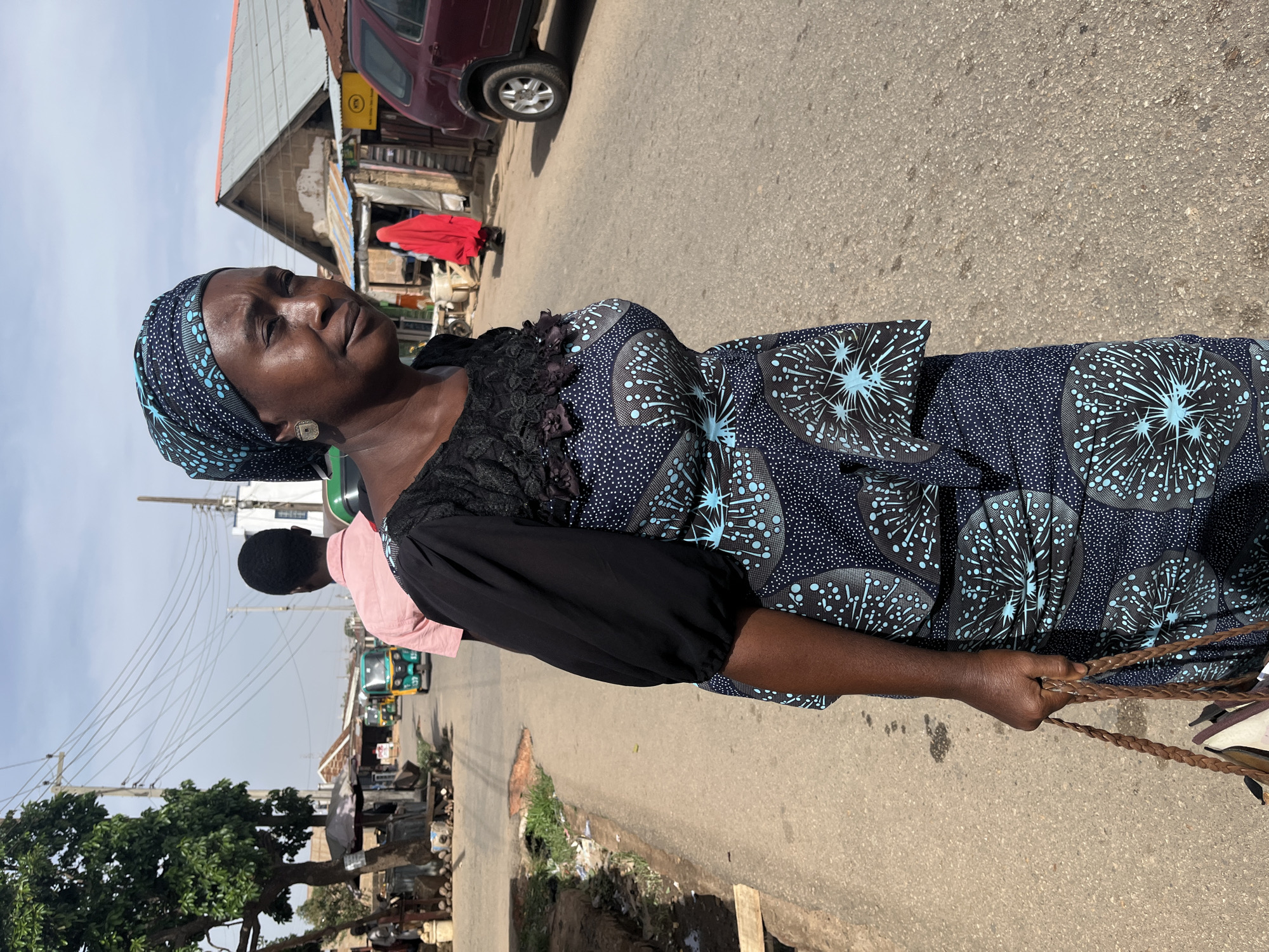 A woman stands on a street in Jos, Nigeria. She is wearing a patterned blue dress and headscarf, with black lace detailing around the neckline and black sleeves. She holds braided straps. The street is paved and appears to be a commercial area with shops and vehicles visible in the background. Other individuals and vehicles are present on the street. A burgundy vehicle is parked near a building on the left. There are power lines and a tree on the left side. The sky is overcast.