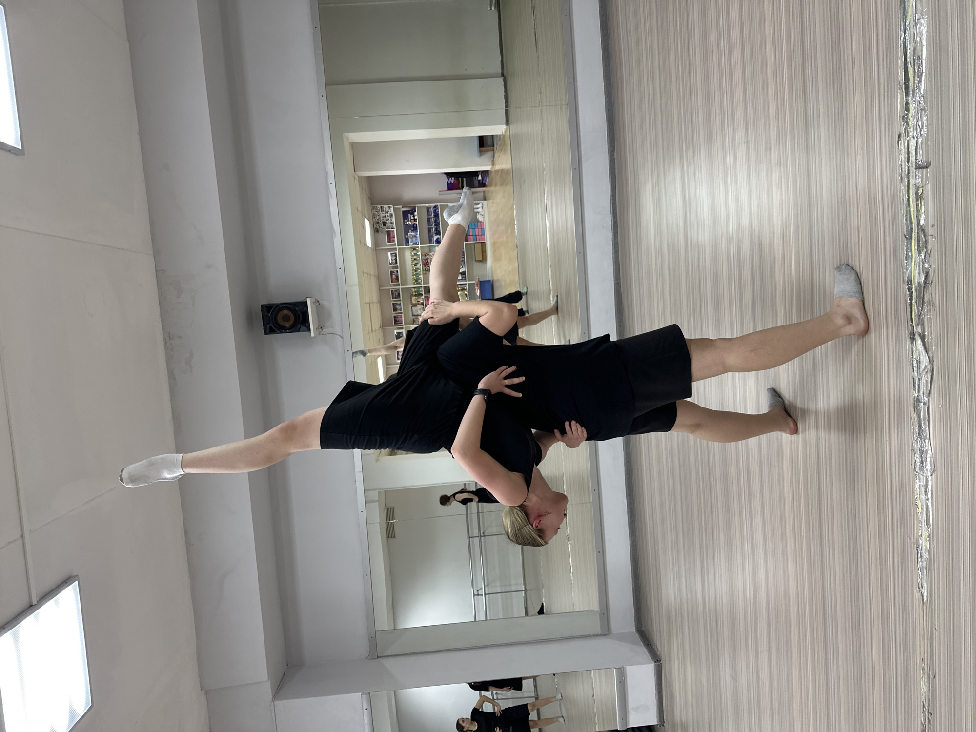 The image depicts an indoor scene within a dance or gymnastics studio, located in the city of Votkinsk, Russia. Two individuals, both adult females, are performing a partner acrobatic lift in the foreground.
The first individual, serving as the base, has light blonde hair and is wearing a black short-sleeved top, black shorts, and grey socks. She supports the second individual on her left shoulder and with both arms, which are wrapped around the second individual's torso and thigh. She wears a dark-colored fitness tracker on her left wrist and her head is tilted downwards.
The second individual, being lifted, is also wearing a black short-sleeved top, black shorts, and white socks. Her body is positioned horizontally, with her right leg extended vertically upwards. Her left leg is bent at the knee, with the foot near her right thigh. Her right arm is extended forward, while her left arm is bent.
The environment features a light-colored, horizontally striped laminate floor. The walls are white, with large mirrors covering a significant portion of the wall behind the subjects. These mirrors reflect other individuals, a ballet barre, and a multi-shelved unit containing various objects, possibly trophies, in an adjacent room. The ceiling is white, equipped with two large rectangular fluorescent light fixtures. A black, square-shaped speaker with a circular element is mounted on the upper wall to the right. A section of metallic-looking tape or worn material is visible horizontally across the floor in the bottom foreground.