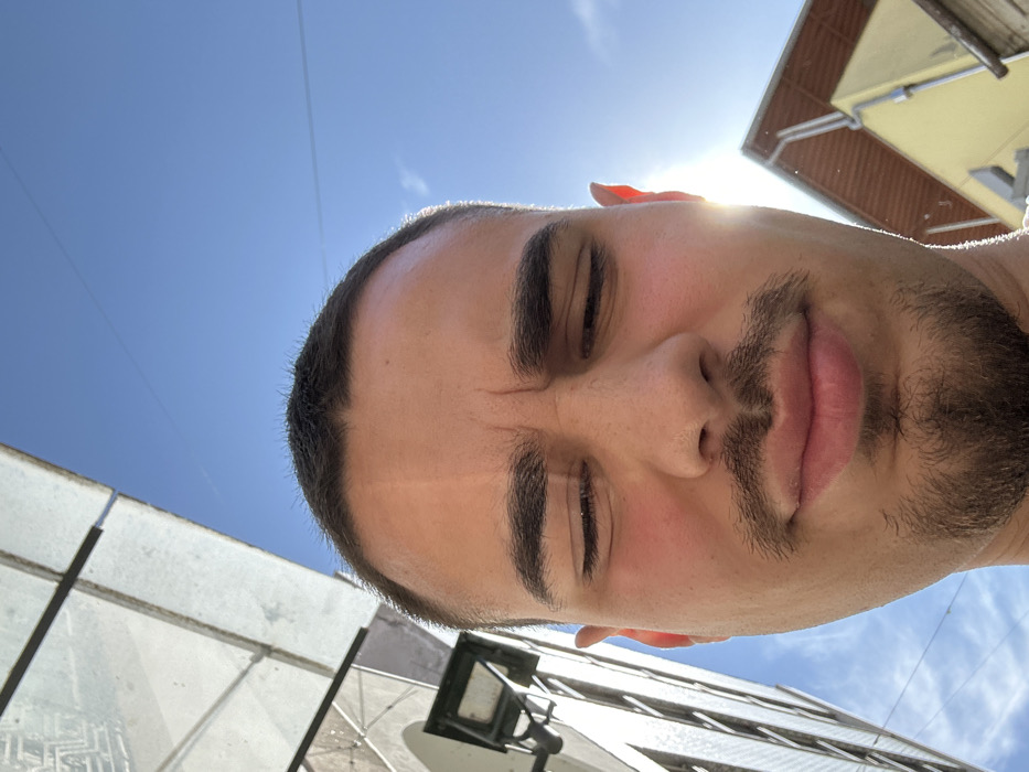 A low-angle close-up photograph captures a male individual with short dark hair, dark eyebrows, a mustache, and a goatee. His eyes are squinted shut, and his forehead is furrowed, suggesting a reaction to bright light. His cheeks exhibit a reddish flush. Strong direct sunlight, visible as a glare, originates from the lower right of the frame, illuminating the subject's right side. The background features a clear blue sky. To the left, a multi-story building with reflective surfaces or windows and an attached street lamp under a glass canopy structure is partially visible. To the right, a yellow-facade building with a brown-tiled roof and a gutter system is observed. Faint utility lines traverse the upper sky. The scene is situated in the city of Chaves, Portugal.