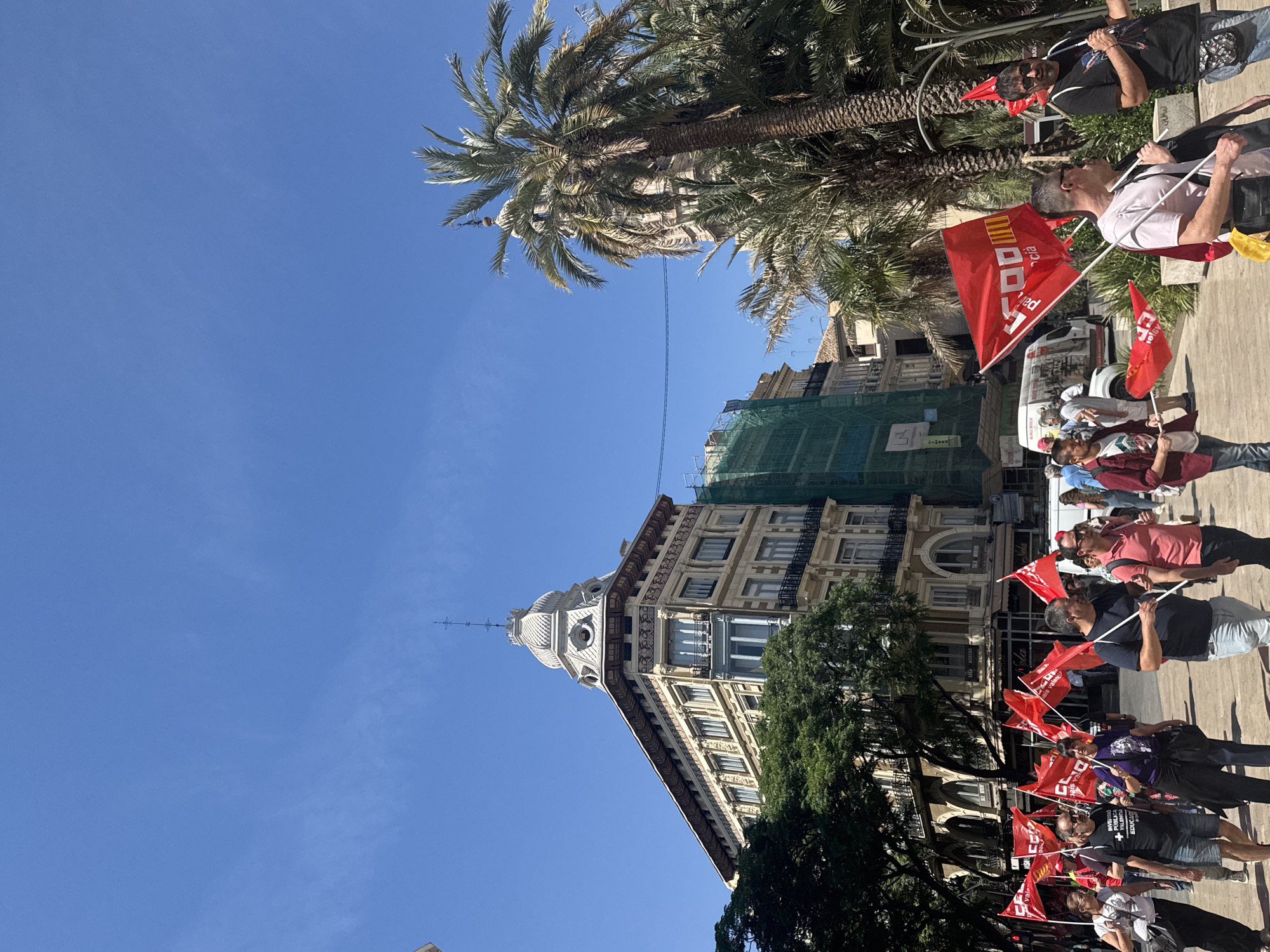 A group of people marches with red flags along a city street in València, Spain. A distinctive wedge-shaped building stands alongside the demonstrators. The building's façade is ornate, with architectural details including a small dome or cupola on its corner. Scaffolding and green netting partially obscures the top section of the building. Palm trees are visible above and to the side of the building, and the sky is clear and blue. The people are walking along a paved area, and some carry or wave red flags. The flags feature writing and symbols. The street appears to be relatively busy, and there is a vehicle in the background. The scene has the characteristics of a protest or demonstration.