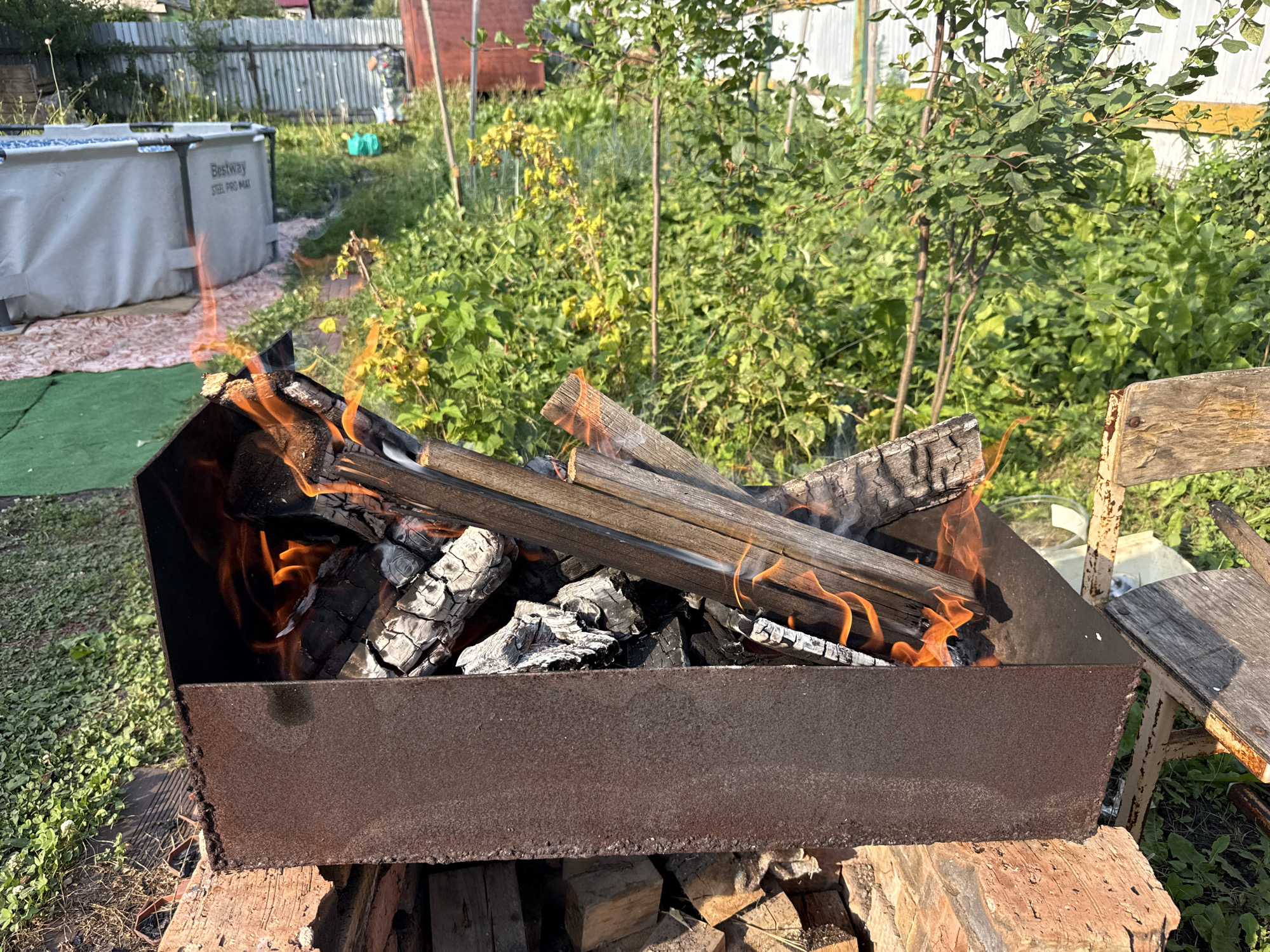 This image depicts a rustic outdoor scene with a fire burning in a metal brazier. The brazier is filled with glowing embers and several pieces of firewood, with flames licking at the wood. It sits on top of a stack of bricks and other wooden materials.
In the background, a weathered wooden chair is visible to the right, suggesting a place for someone to sit and enjoy the fire. Beyond that, lush green foliage and trees create a natural backdrop. To the left of the brazier, a large, grey inflatable swimming pool with "Bestway STEEL PRO MAX" text is partially visible. A green mat lies in front of it. The overall setting appears to be a backyard or garden area, possibly a dacha in a rural or suburban location.
The sunlight suggests it's daytime, likely afternoon, given the warm tones of the fire and the shadows. The weather seems pleasant and conducive to outdoor activities. There are no people visible in the image, focusing the viewer's attention on the fire and its surroundings. The scene evokes a sense of warmth, relaxation, and simple outdoor enjoyment.