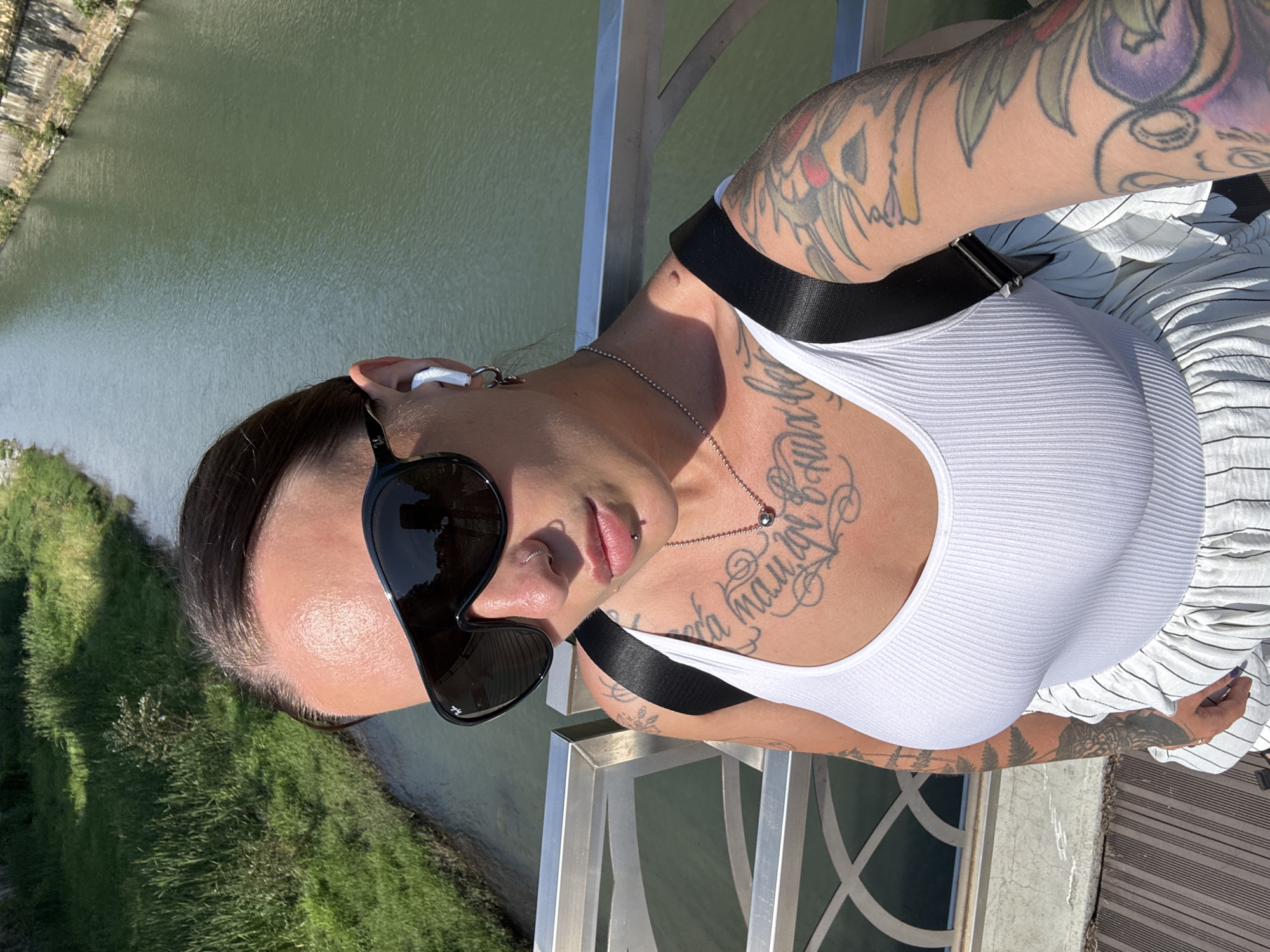 A woman with dark hair and tattoos on her arms and torso is shown in a close-up, portrait-style image. She is wearing a white, ribbed tank top and sunglasses. Her left ear has a white earbud. She is also wearing a silver chain necklace with a small circular pendant. She has a nose ring and a lip piercing. The background shows a body of green water, possibly a river or canal, with a grassy bank on the left side. A metal railing is visible in the foreground, suggesting she is on a bridge or elevated walkway. The lighting indicates it is daytime, likely sunny due to the reflection in her sunglasses. The overall mood is casual and relaxed.