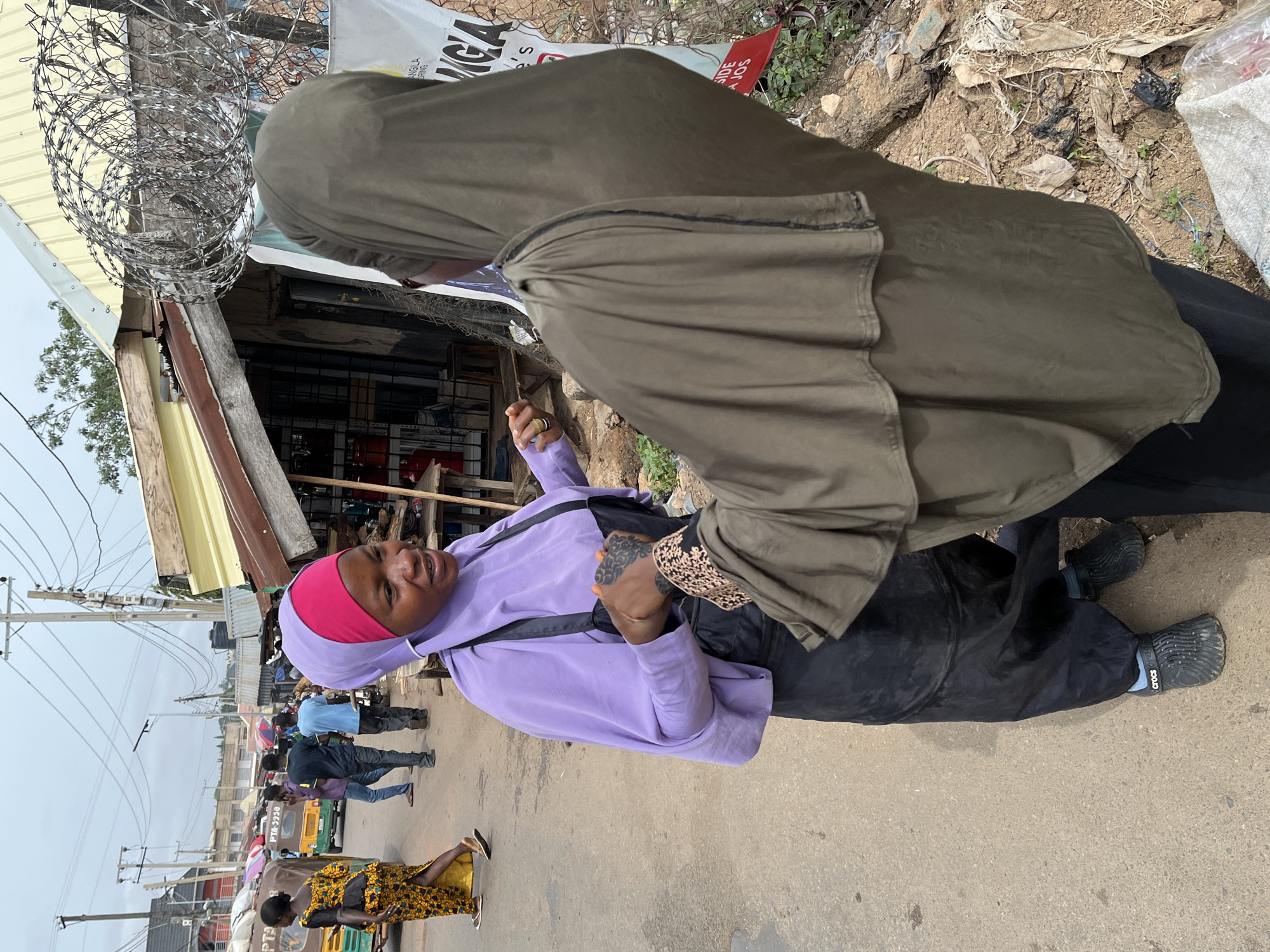 The image captures a street scene in Jos, Nigeria. Two women, both wearing hijabs, are the central focus. One woman wears a purple hijab and lavender blouse, and the other wears an olive green hijab. They are standing near a building with barbed wire on its roof. Other people are seen walking on the street in the background. There are utility poles and various commercial establishments along the street.
