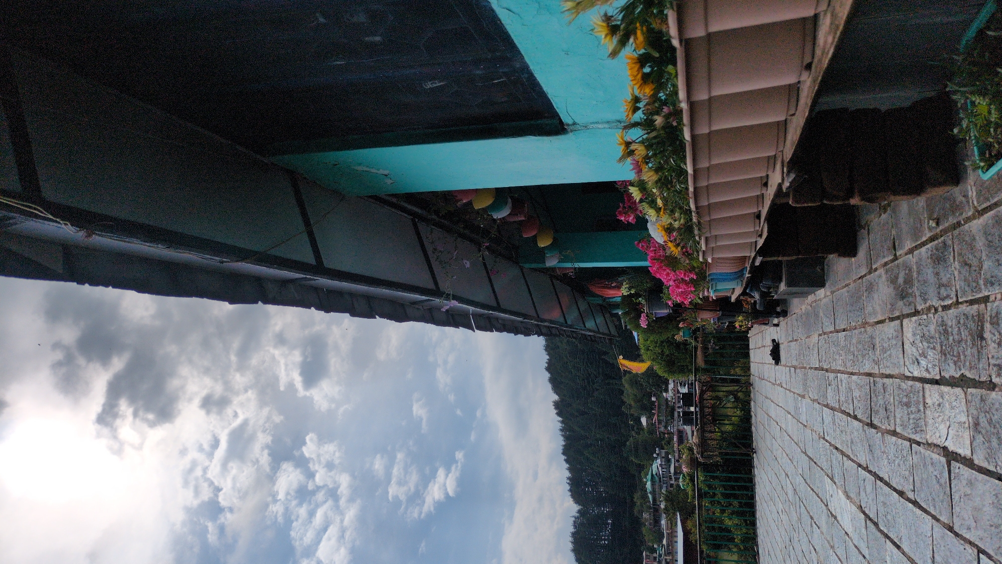 The image presents an outdoor scene, seemingly shot in the city of Haini, India. The perspective is upward-angled, showcasing a view from under a covered structure. To the left, a dark, possibly metal or tiled awning is visible against a cloudy sky. Progressing rightwards, a teal-painted structure appears, adorned with hanging, colorful, teardrop-shaped decorations.
A flower-filled planter box runs horizontally along the scene, featuring a mix of yellow, pink, and red blooms. Beyond the planter box, a green, tree-covered landscape stretches towards the horizon, interspersed with buildings.
In the foreground, a gray, tiled or bricked pathway extends towards the viewer. On the pathway, two people are present: one near the flower box and one further down the path. A dark, possibly black, object is present, although its precise nature is unclear.